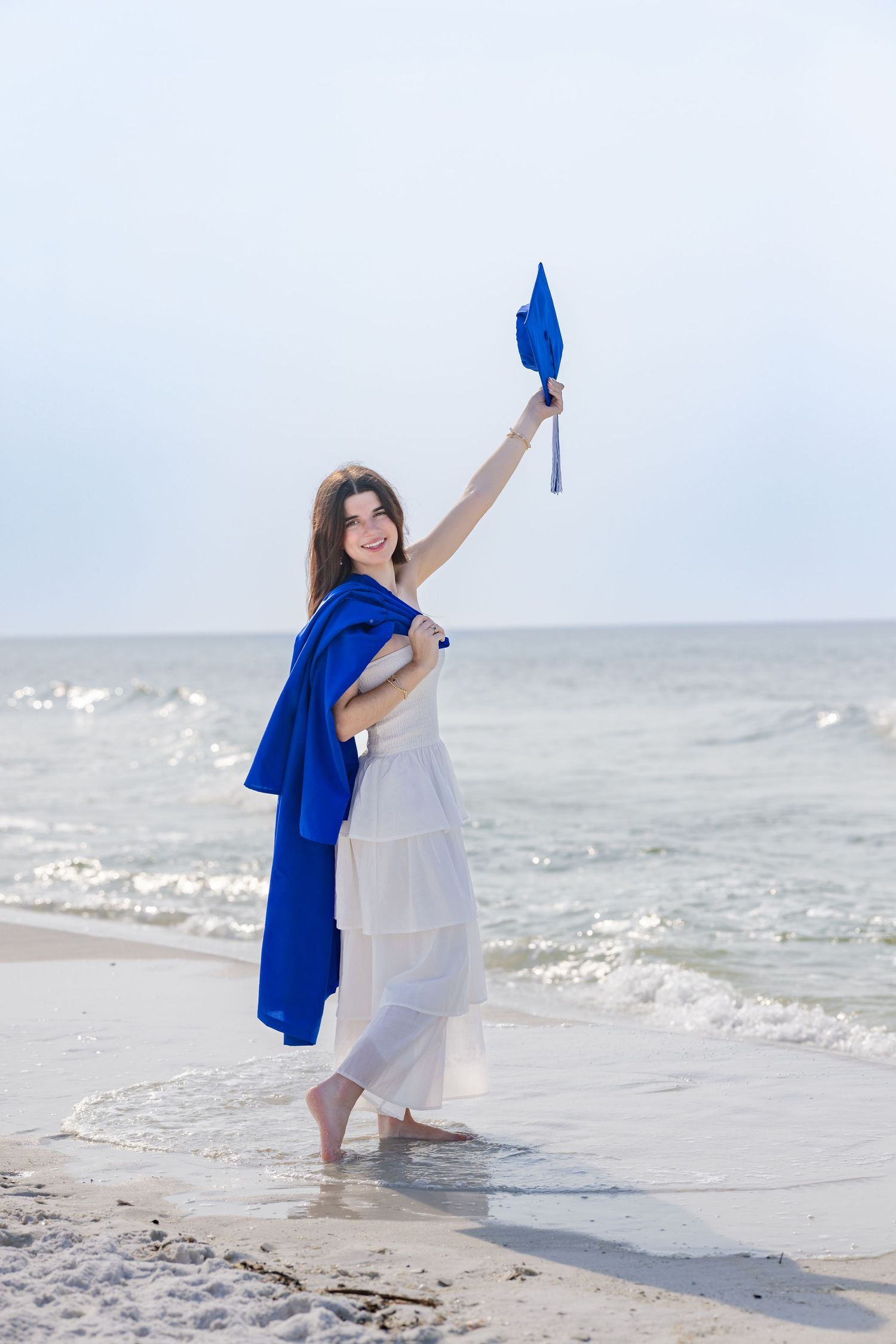 Young woman in white dress holding blue graduation cap on beach, celebrating academic achievement by the ocean.