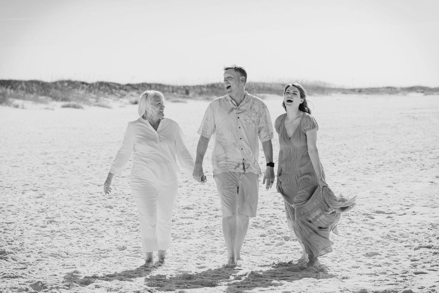 Black and white photo of three people walking and laughing on a sandy beach, dunes in background.