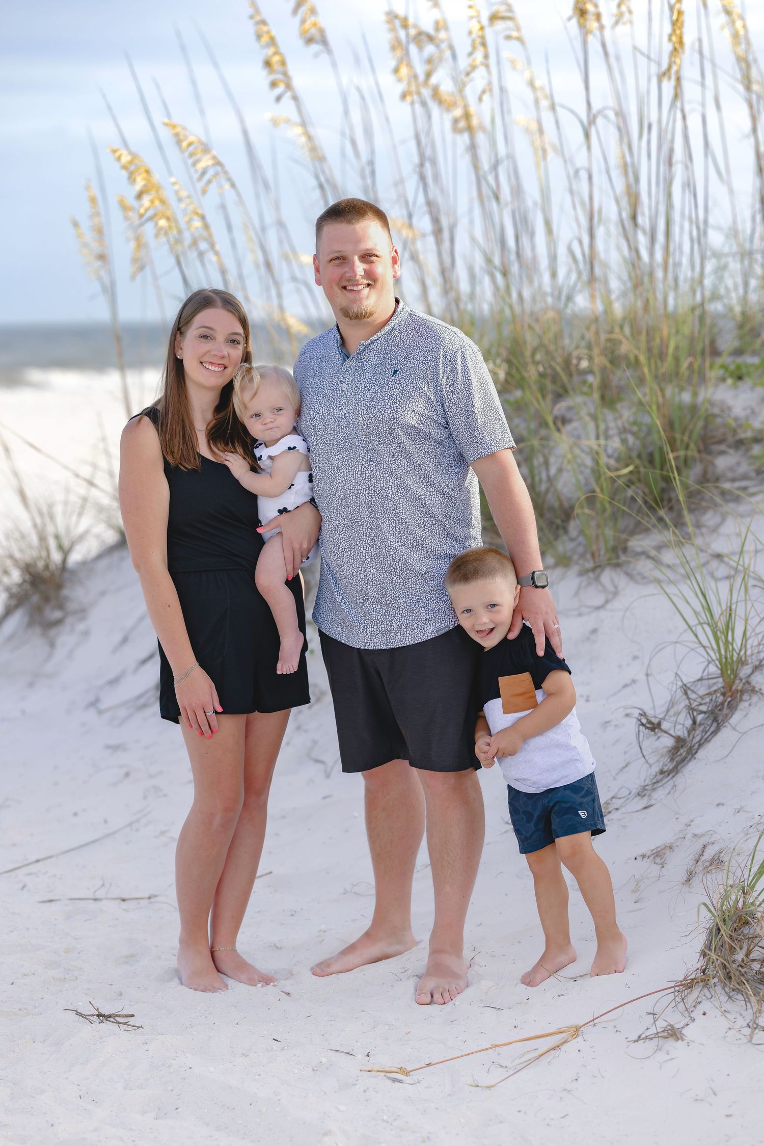 Family of four posing on white sand beach dunes with sea oats, parents with baby and toddler boy.