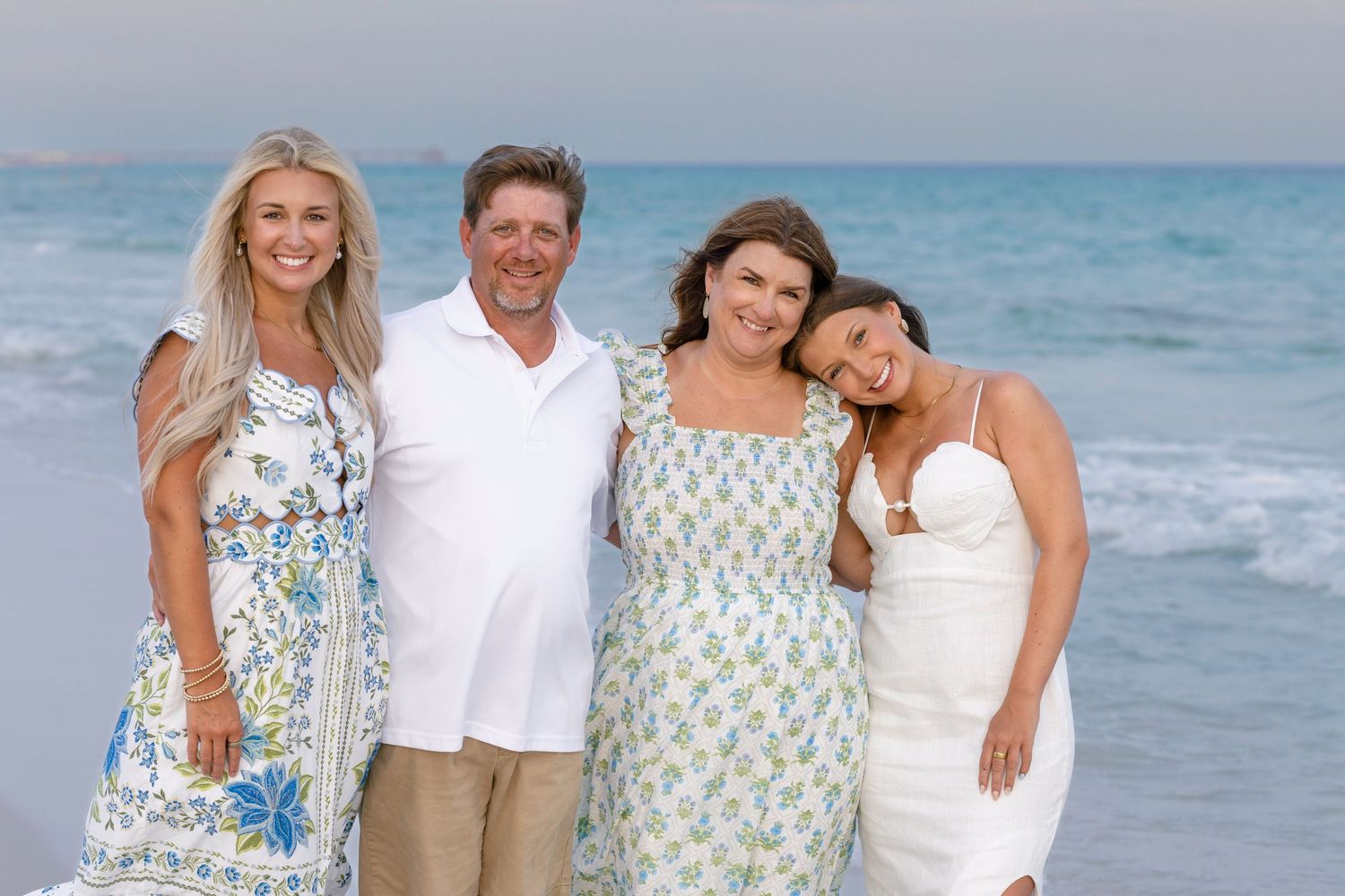 Family of four smiling together on a beach with ocean waves, dressed in summer whites and florals.