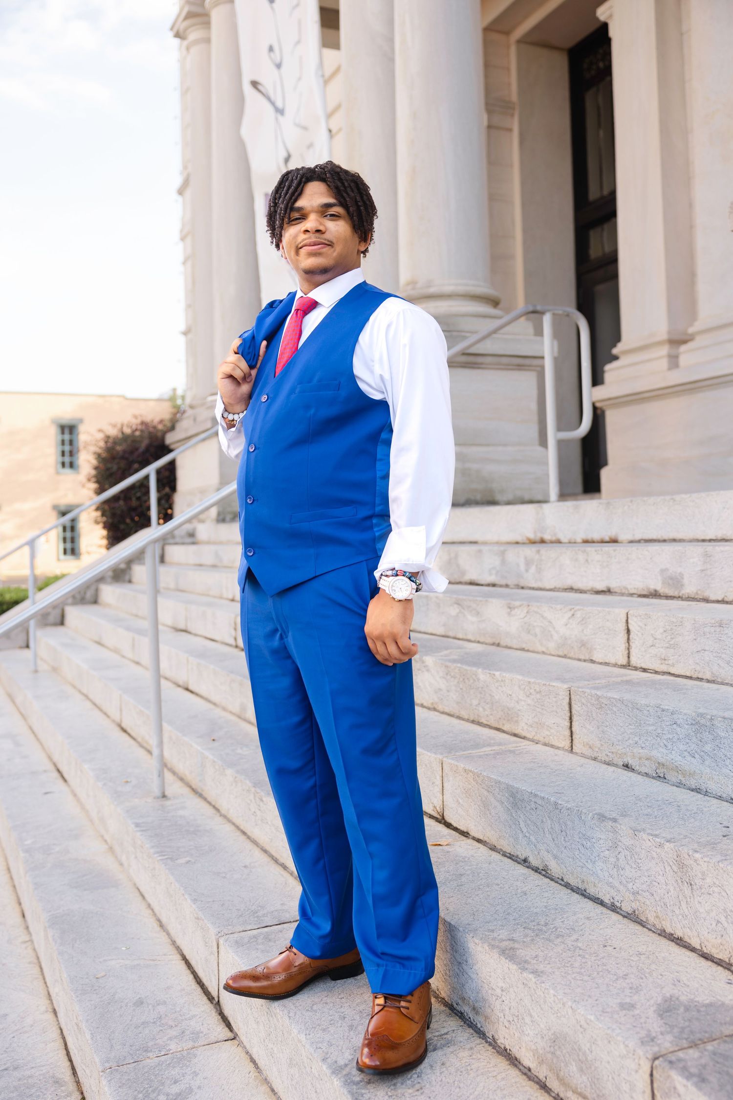 Young man in blue suit vest and pants with red tie poses on stone steps outside a formal building.