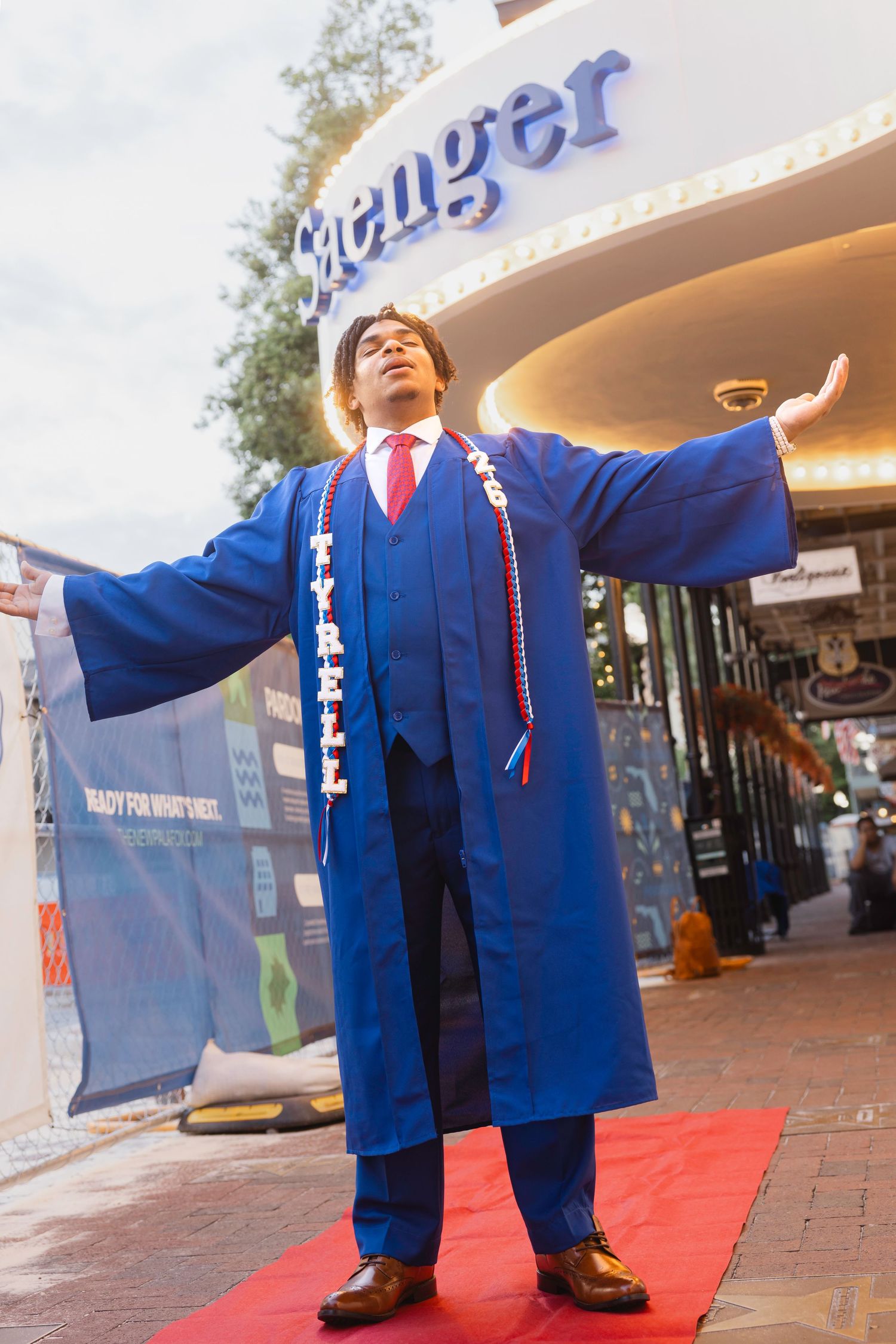 Graduate in blue cap and gown named Tyrell poses with arms outstretched on red carpet outside Saenger Theatre.