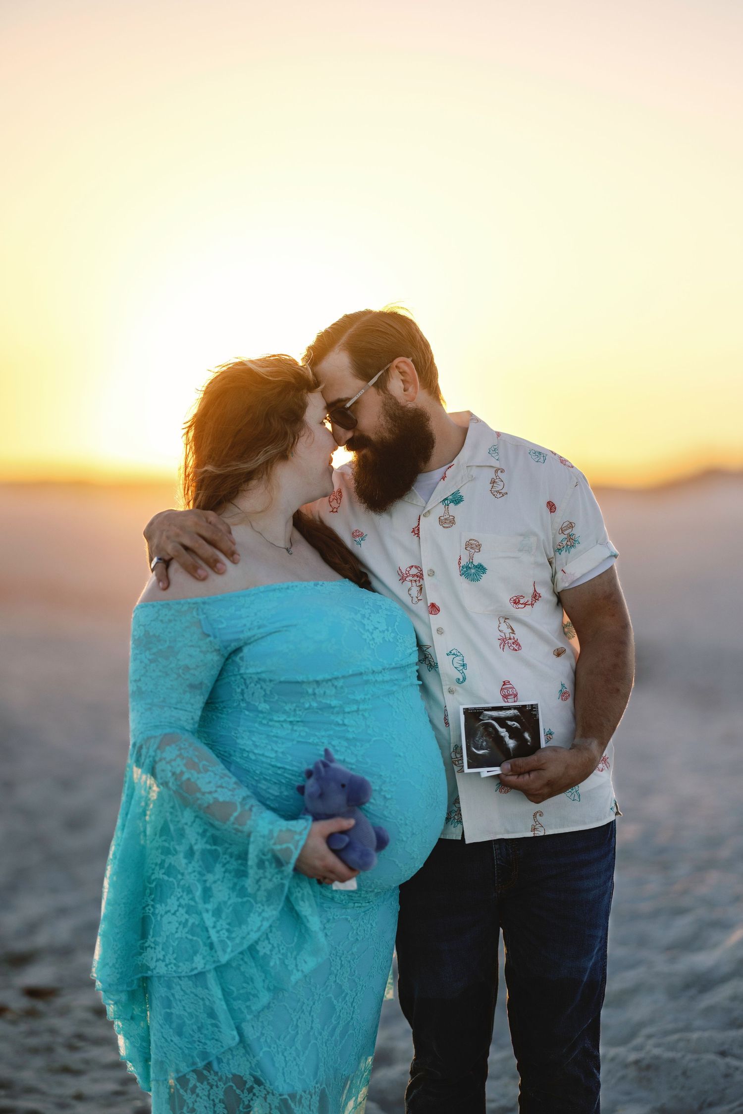 Couple sharing a kiss at sunset on beach, man holding ultrasound photo, woman in teal maternity gown.