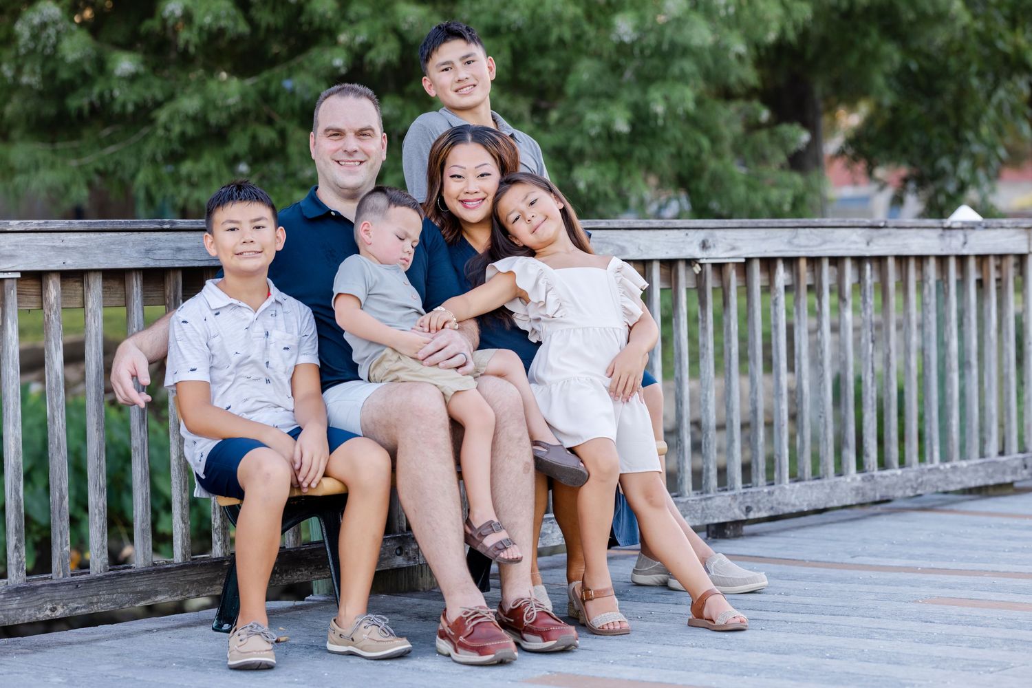 A family sits and smiles together on a wooden deck railing with green foliage in the background.