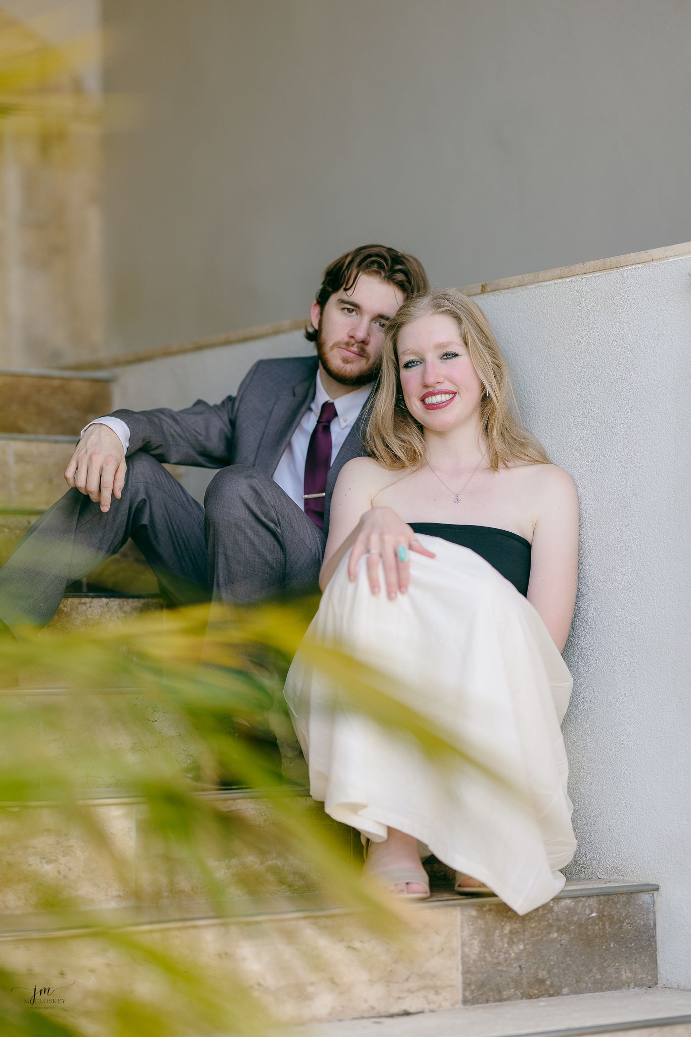 A couple in wedding attire sits together on concrete steps with a dreamy blurred effect in foreground.
