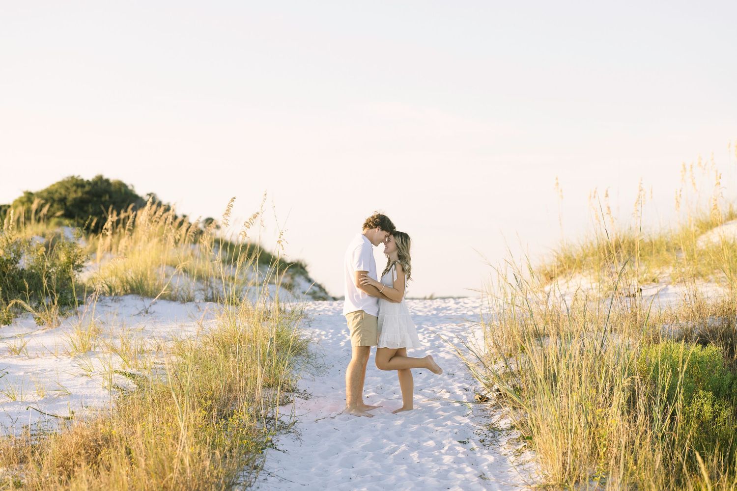 A scenic beachside photoshoot on white sandy dunes with tall grass and golden sunlight filtering through.