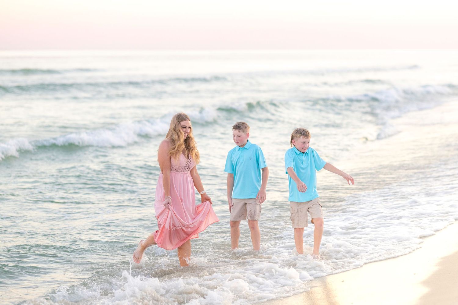 Family walking along the ocean shoreline at sunset in pastel colors.
