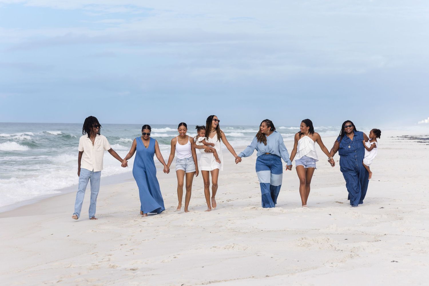 A group of women holding hands and walking together on a white sandy beach with ocean waves in the background.