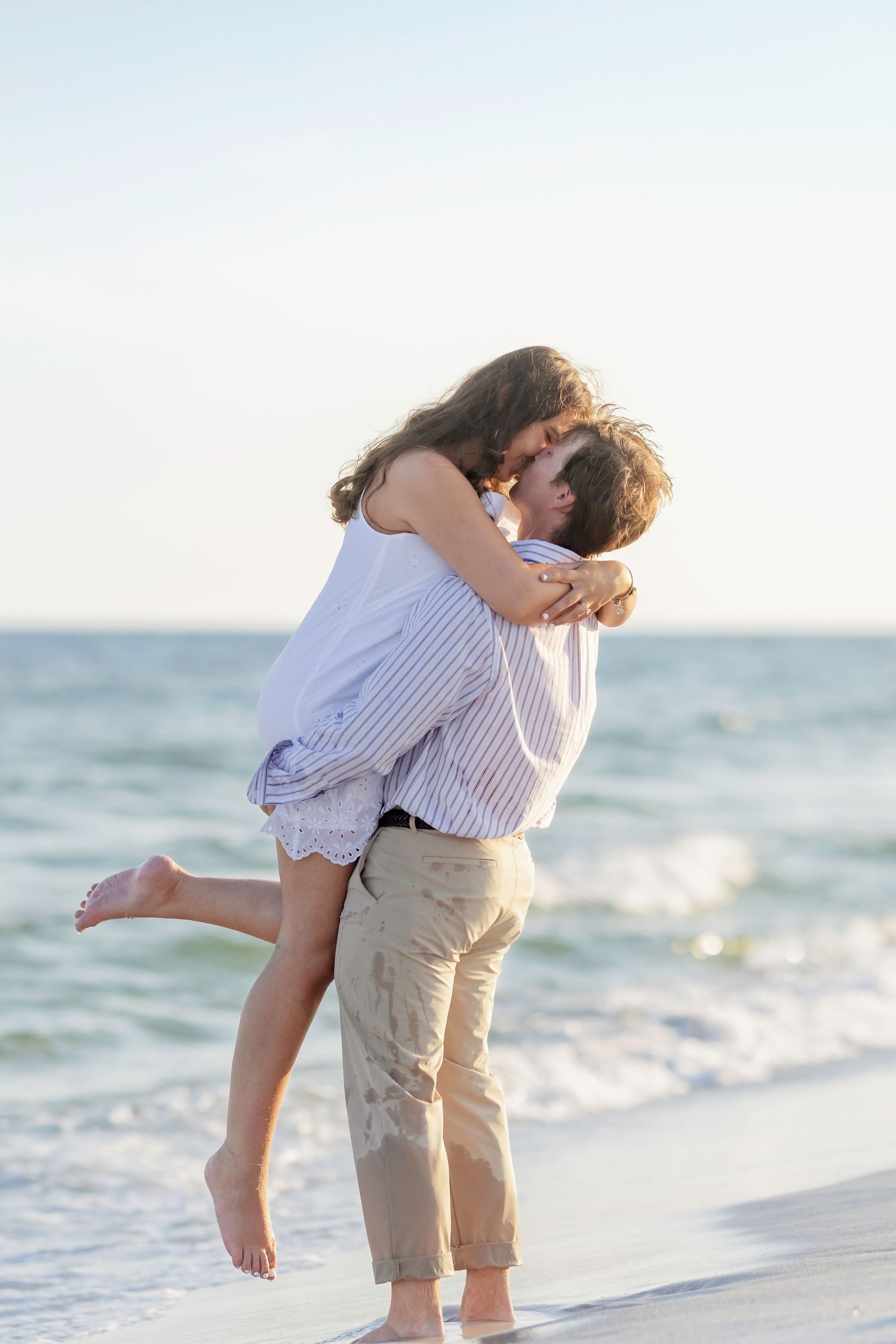 A couple embraces and kisses on a sunny beach with ocean waves in the background.