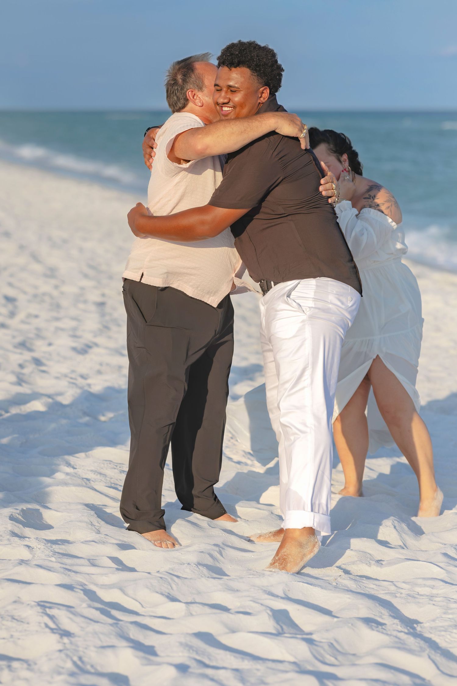 Two couples embrace in a warm group hug on a white sandy beach with calm blue ocean waves in the background.