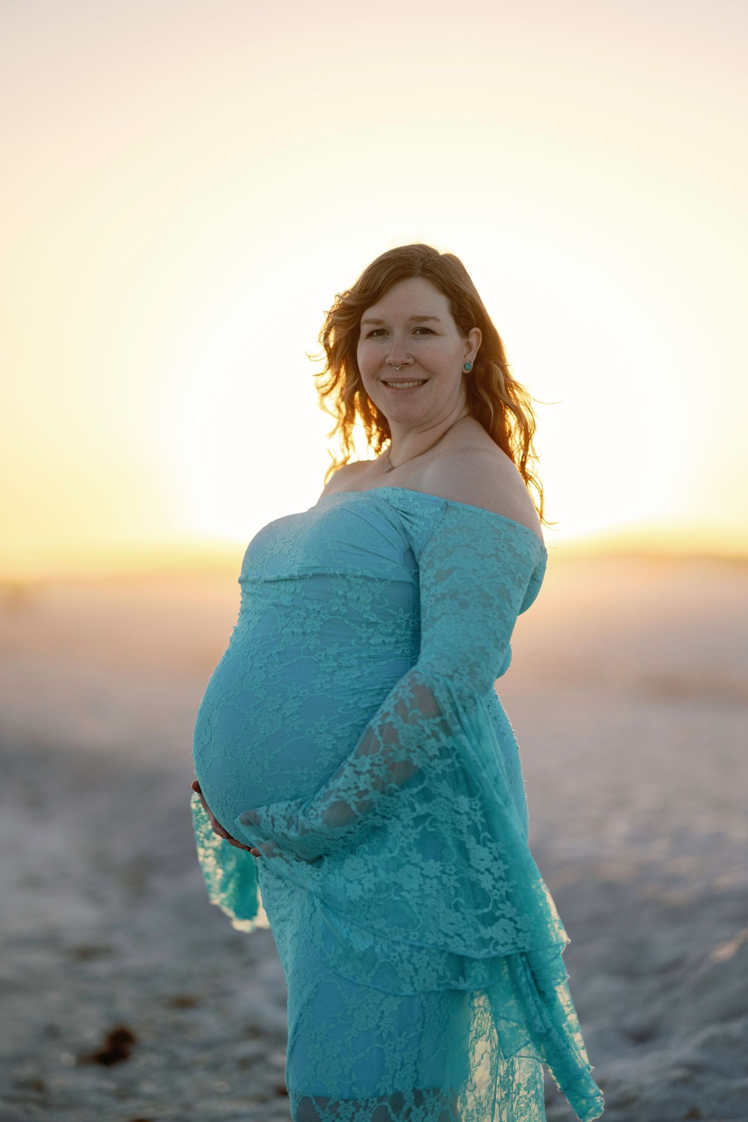 Pregnant woman in teal lace off-shoulder dress smiling at sunset on beach, cradling baby bump.