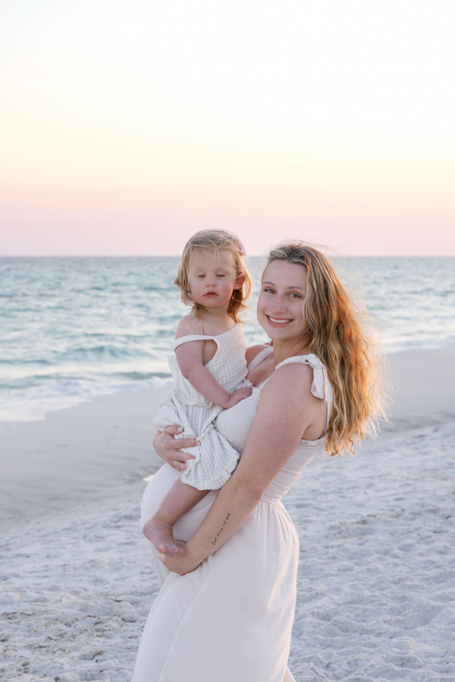 A smiling mother in a white dress holds her young daughter on a serene beach at sunset.