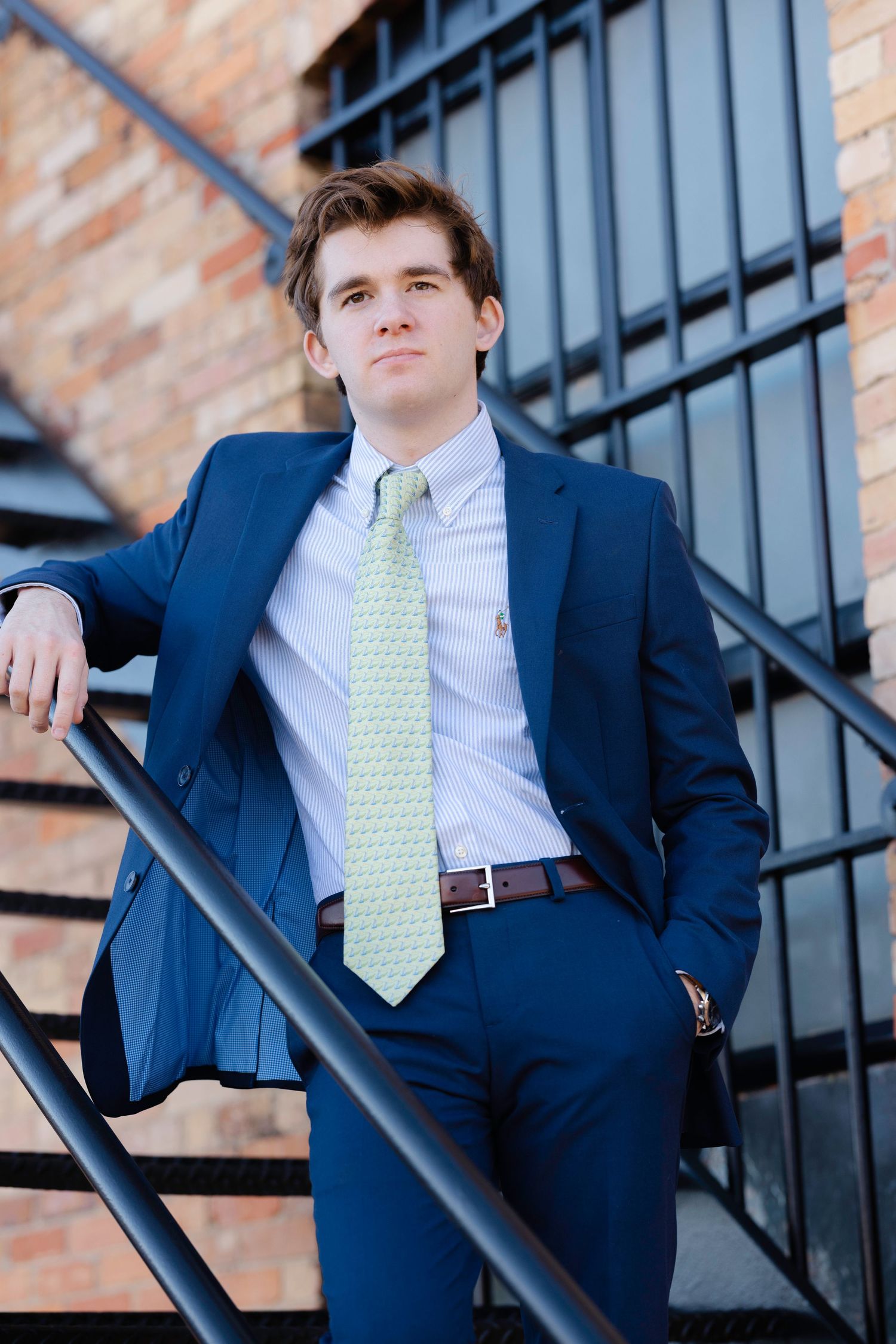 Young man in navy blue suit and green tie poses confidently on outdoor metal staircase against brick wall.