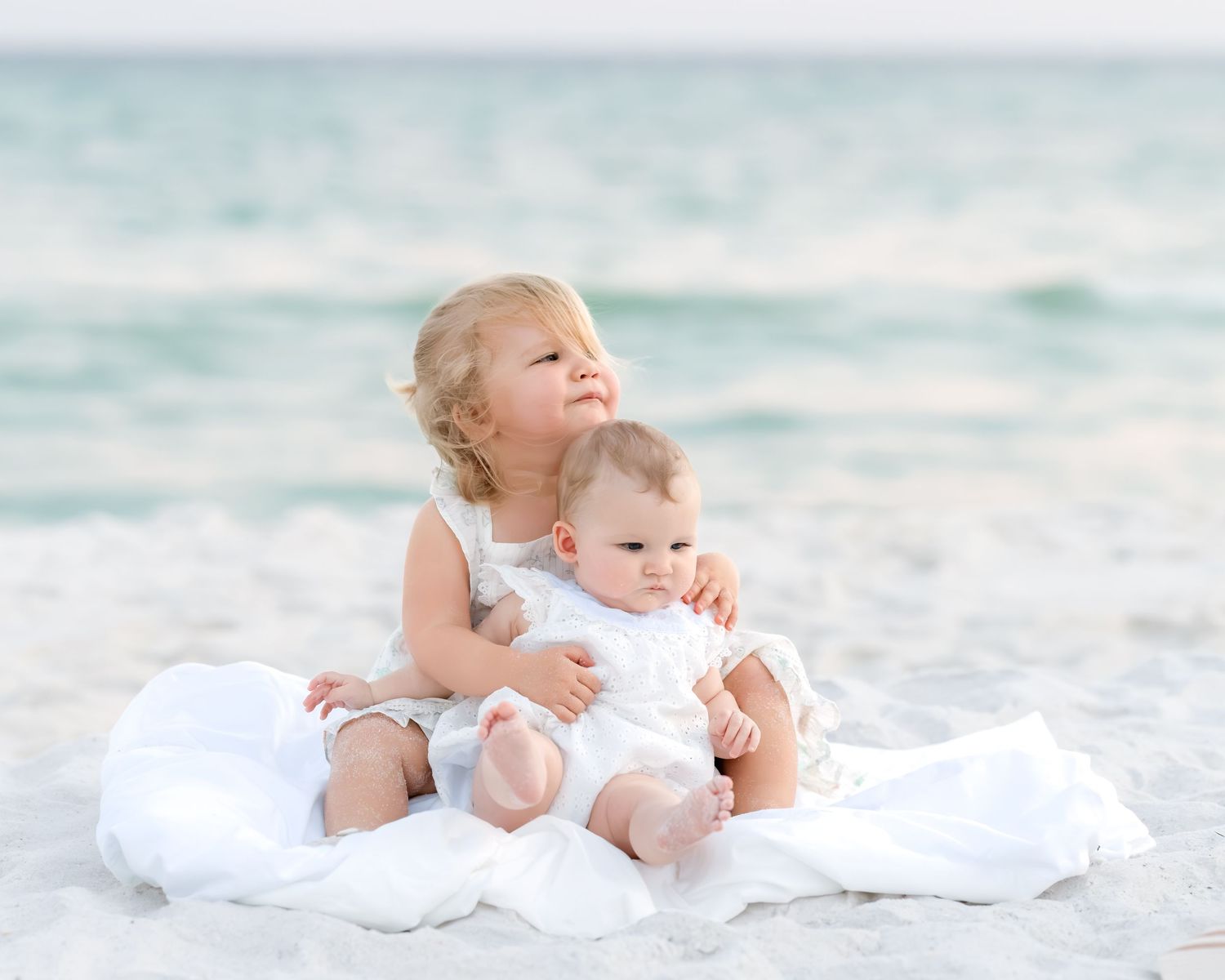 Two young sisters in white dresses sit together on a sandy beach with calm ocean waves in the background.