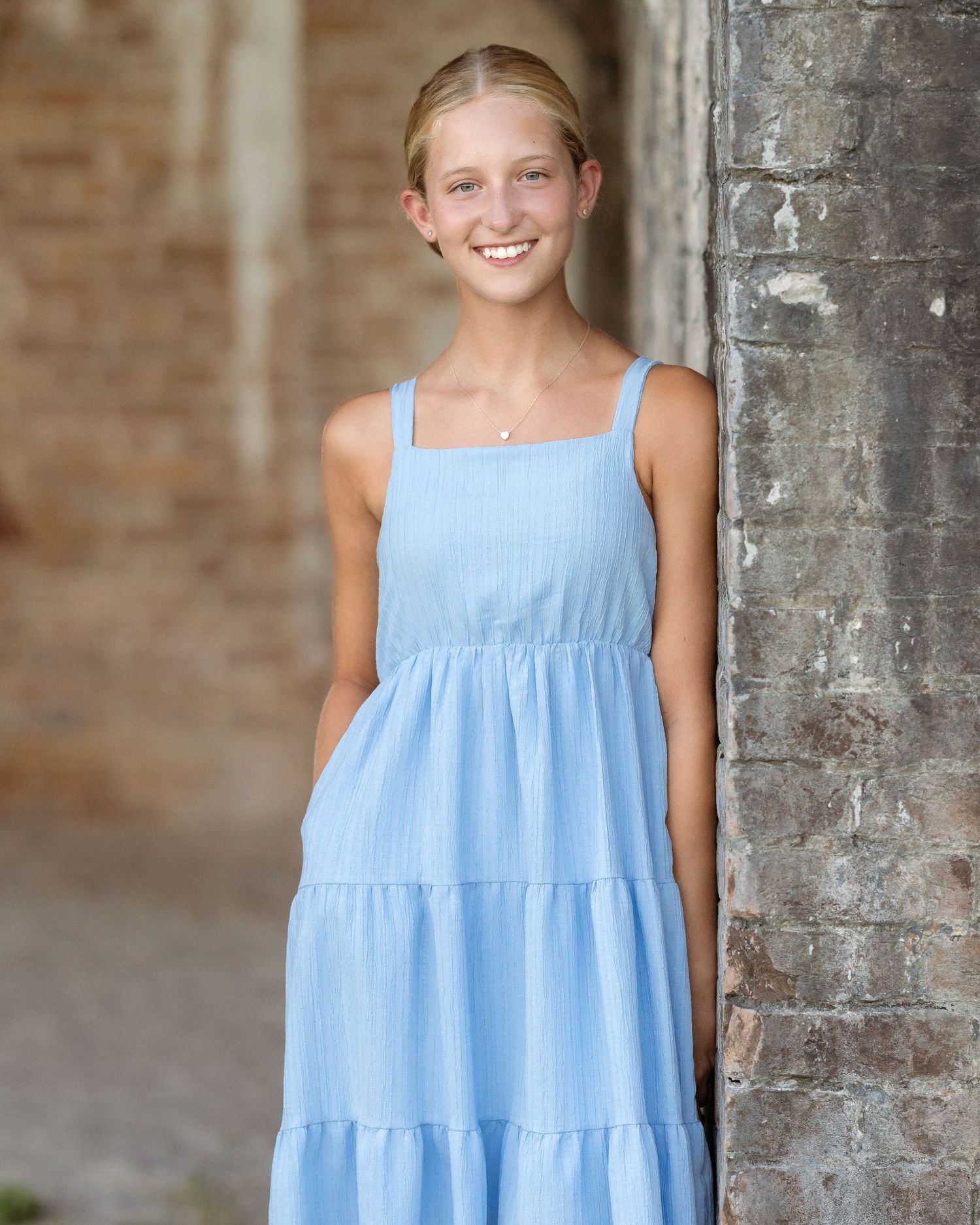 Smiling teenage girl in a light blue tiered sundress leaning against a brick wall outdoors.