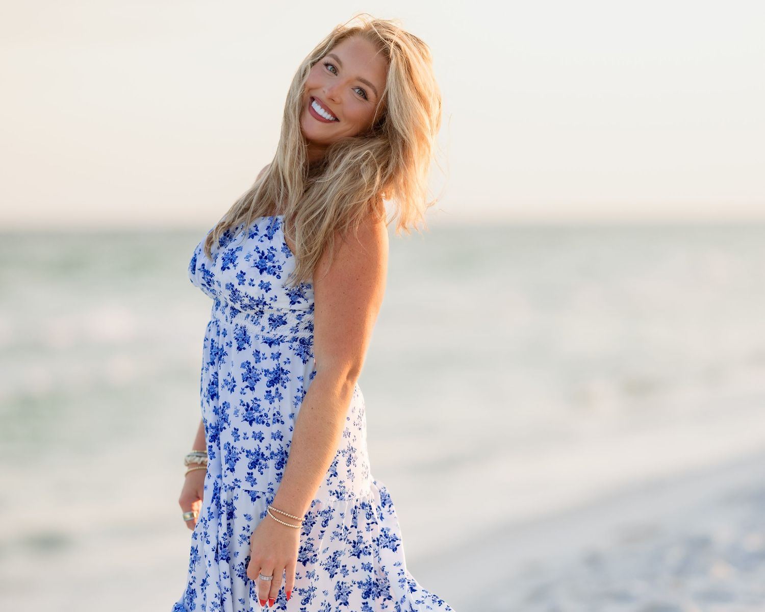 Blonde woman in blue floral sundress laughing joyfully on a beach at golden hour.