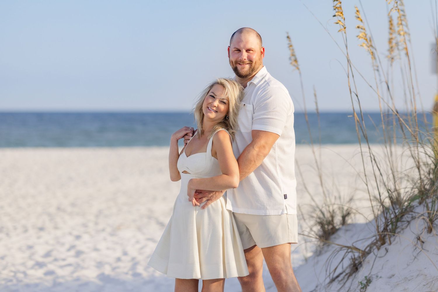 Couple posing together on a white sand beach, woman in white dress and man in white shirt, with sea oats in background.