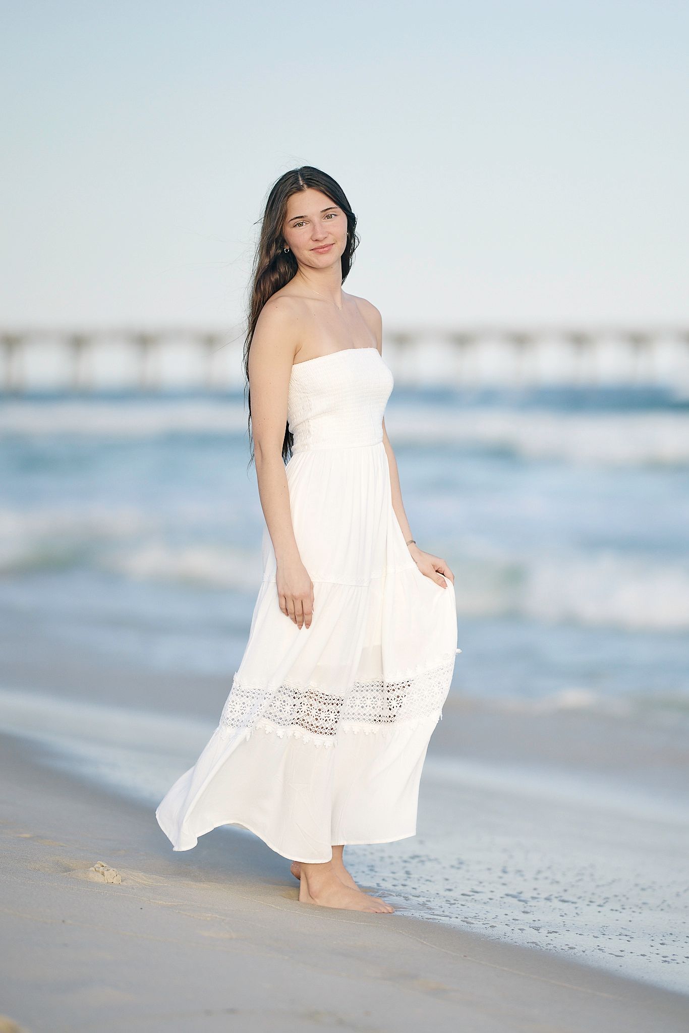A white maxi dress flows in the ocean breeze as waves crash on the beach behind a pier.