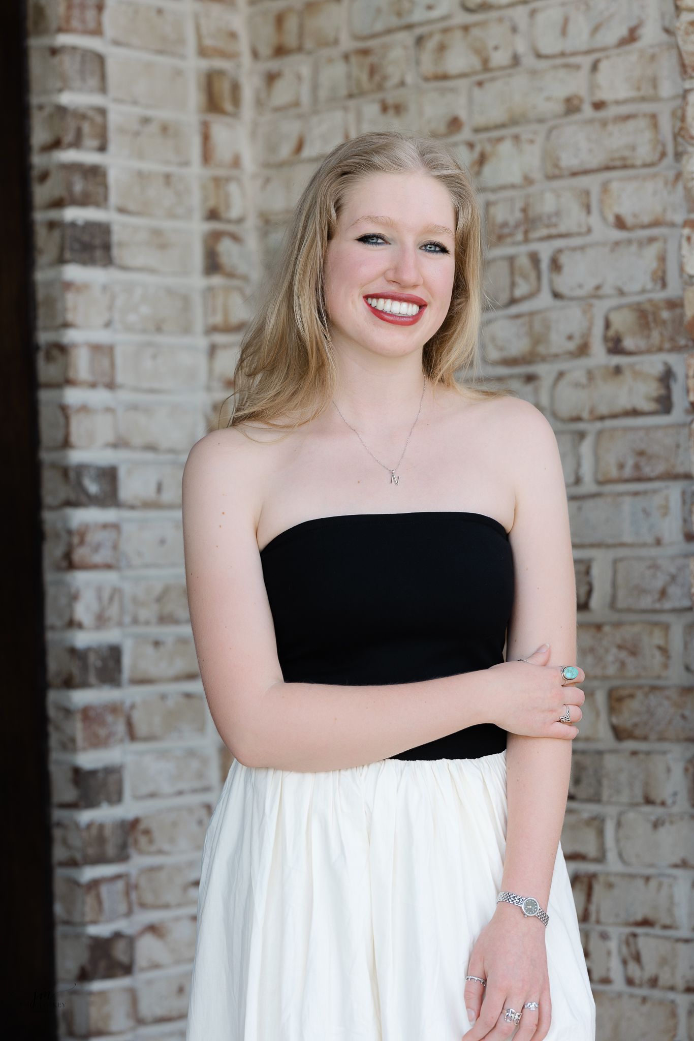 A smiling woman in a black and white strapless dress poses against a brick wall.