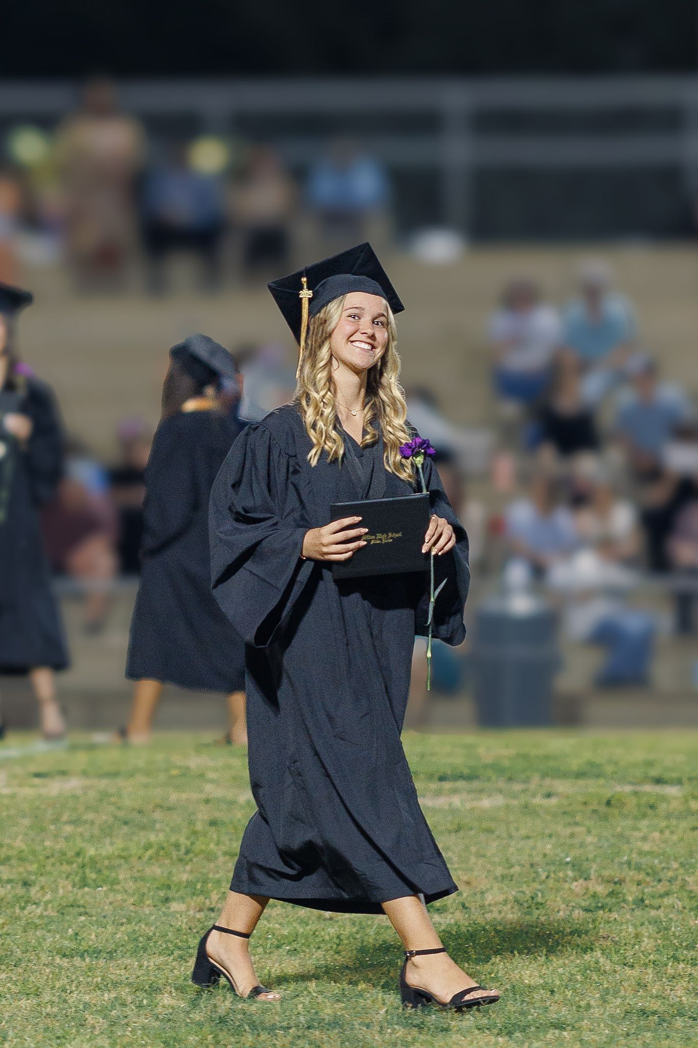 Graduate walks across grass field holding diploma while wearing traditional black cap and gown.