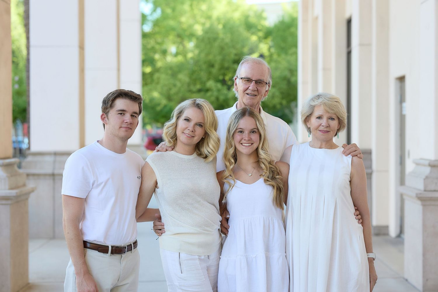 A family of five wearing coordinated white outfits poses together outside a building during a sunny day.