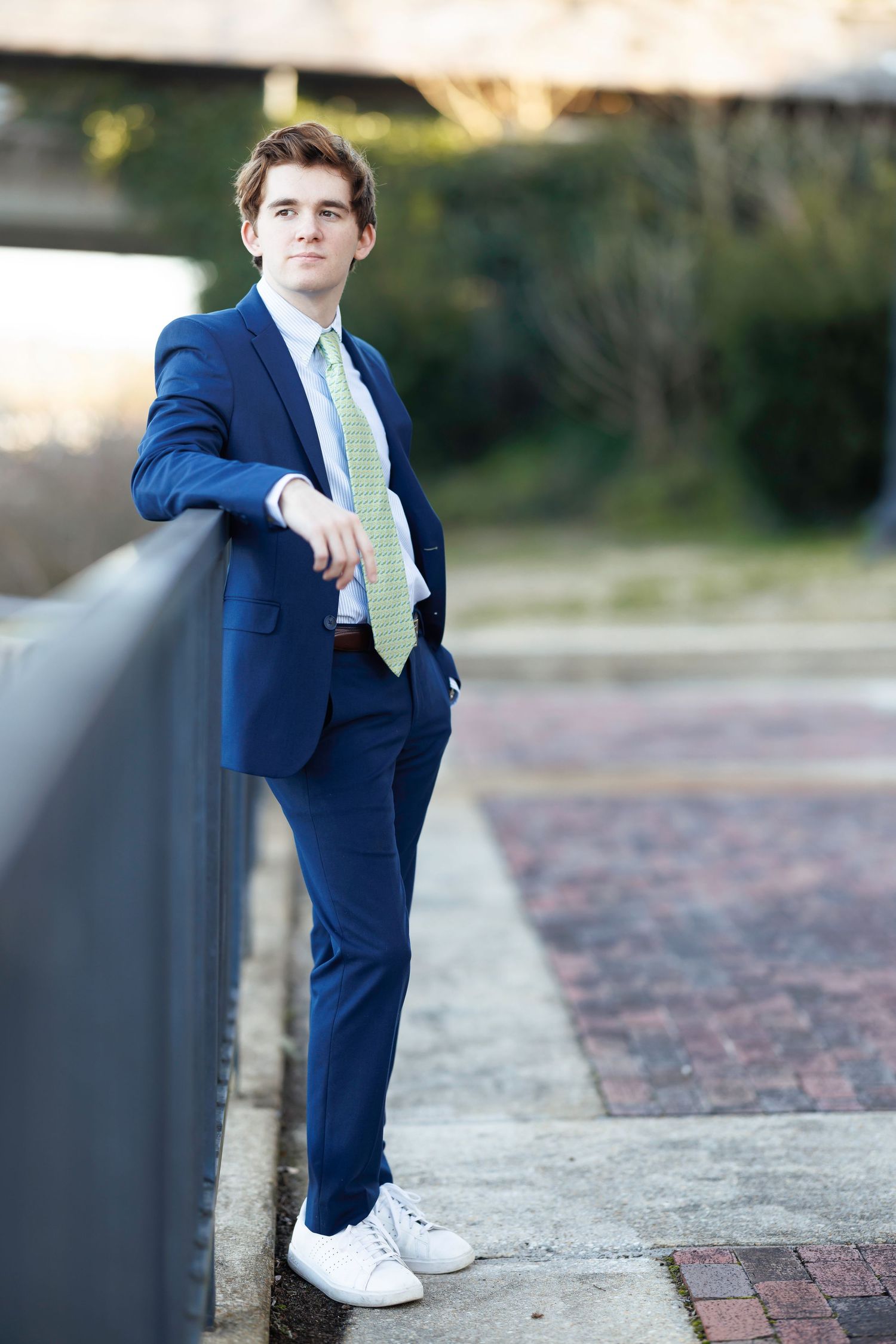 Young man in blue suit, green tie, and white sneakers leaning on railing outdoors in urban setting.