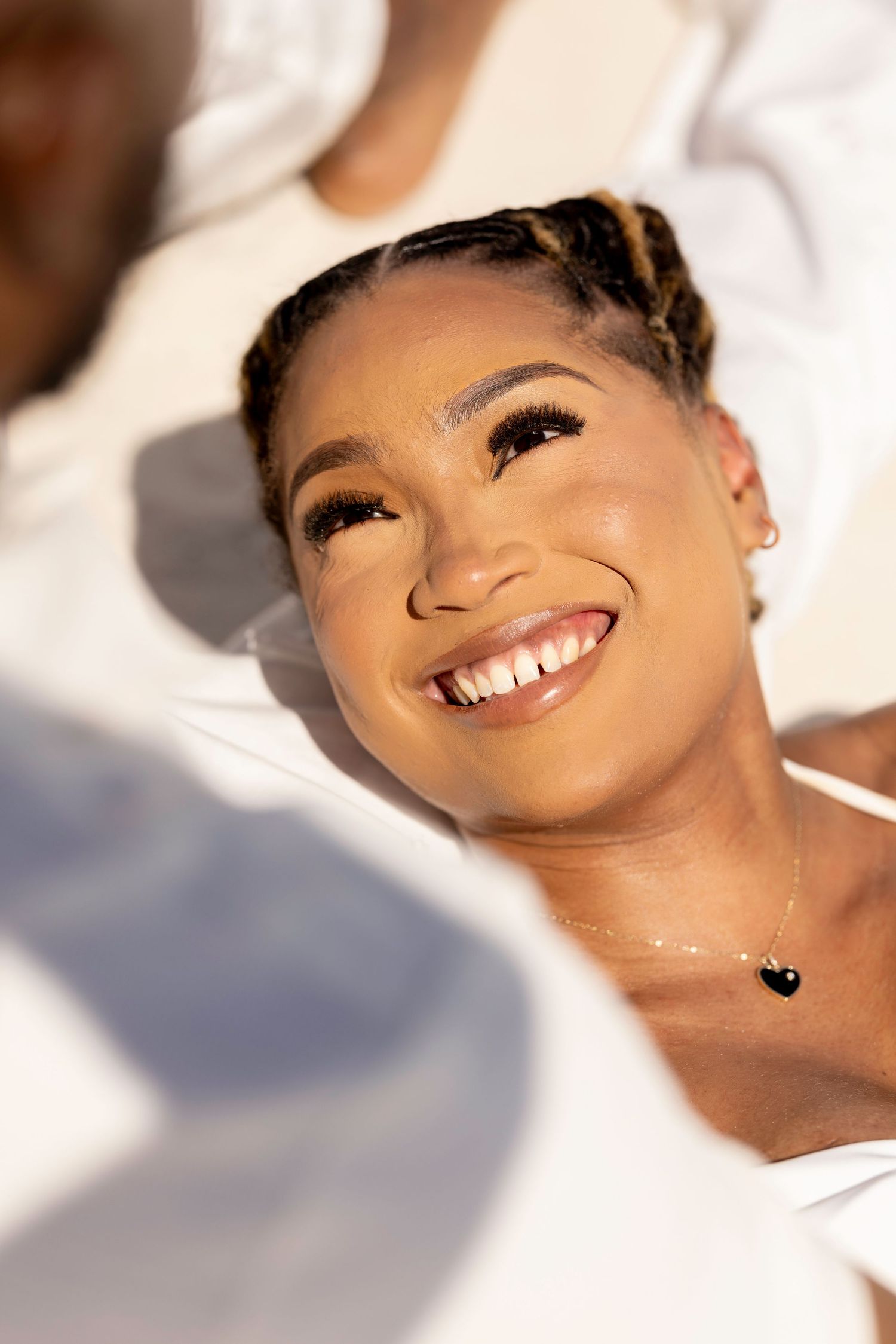 Smiling young woman with braids and gold eyeshadow wearing white, bathed in warm sunlight outdoors.