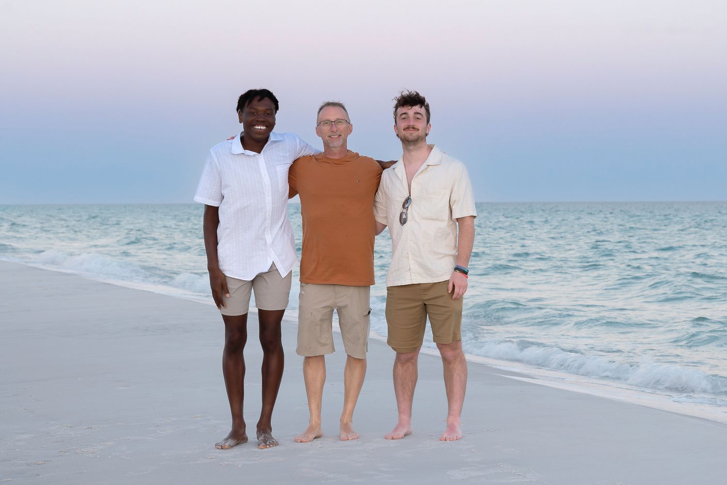 Three men smiling together on a sandy beach at sunset with calm turquoise ocean waves in the background.