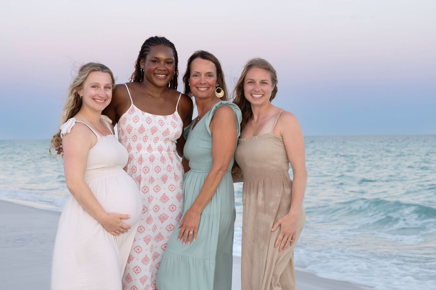 Four women smiling on a beach at sunset, one visibly pregnant, wearing elegant dresses in soft pastel tones.