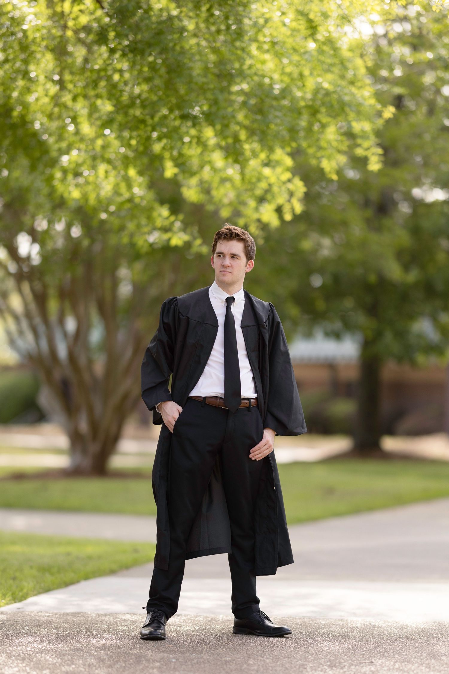 Young man in black graduation gown and tie standing confidently outdoors on a pathway surrounded by green trees.