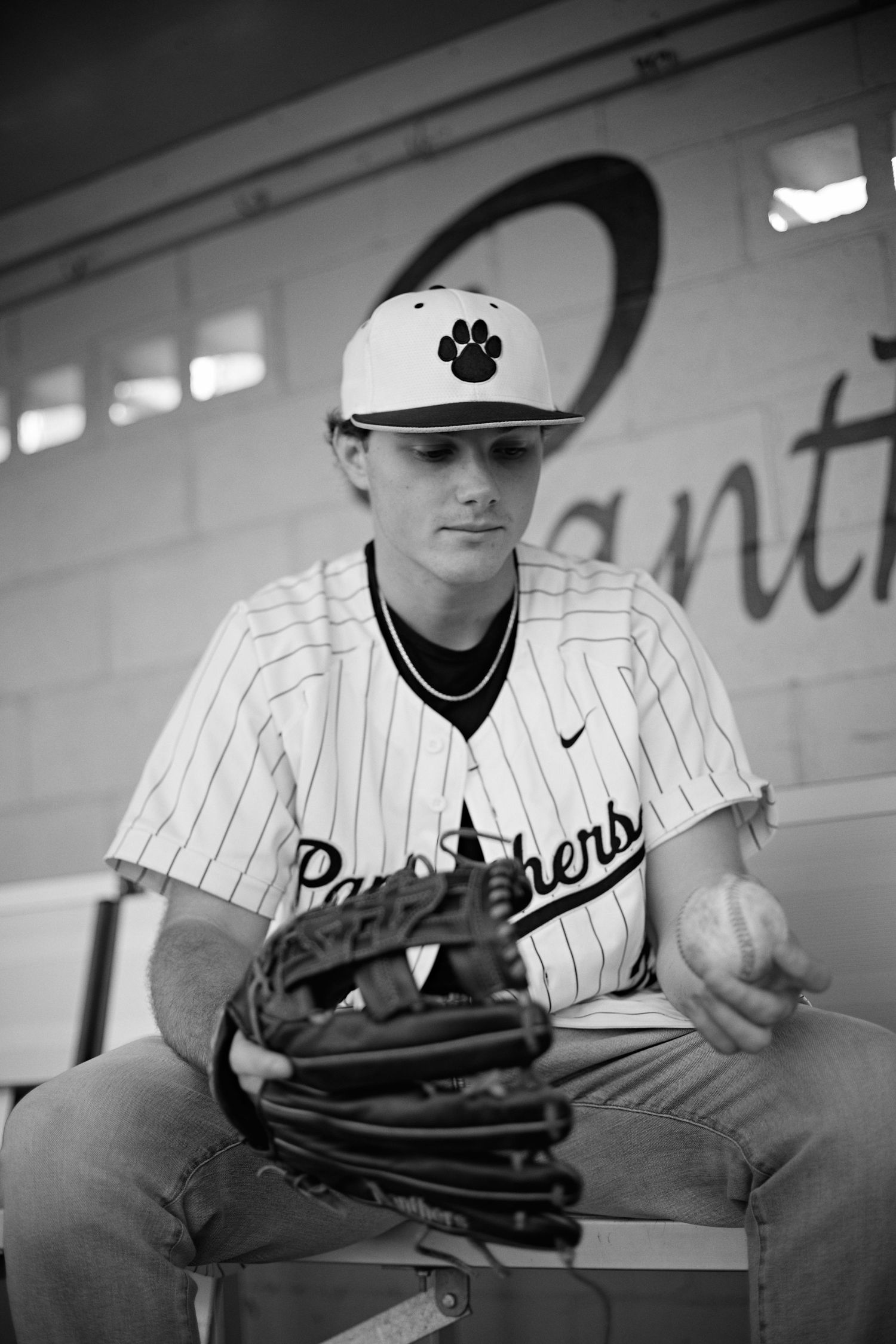 Black and white photo of a baseball player in pinstripes holding a glove and ball in the dugout.