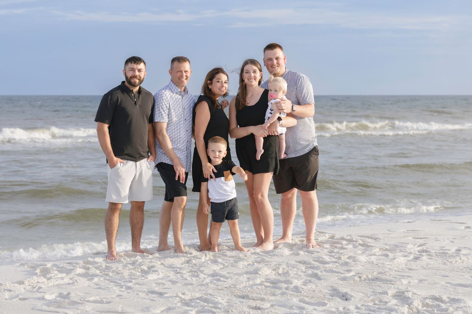 Family of six posing together on a sunny beach with ocean waves in the background.