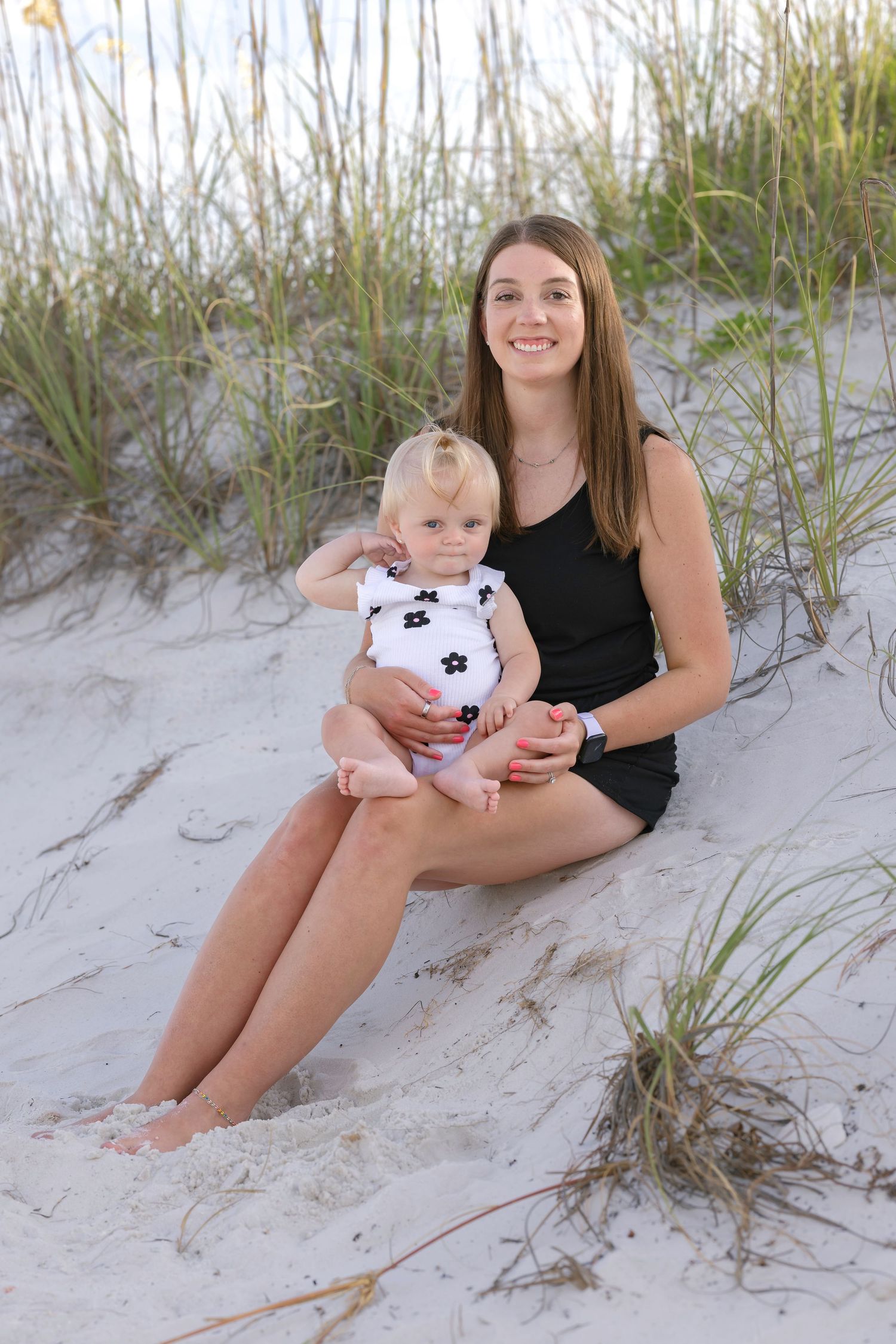 Young mother sitting on sandy beach dunes holding smiling baby girl in floral swimsuit.