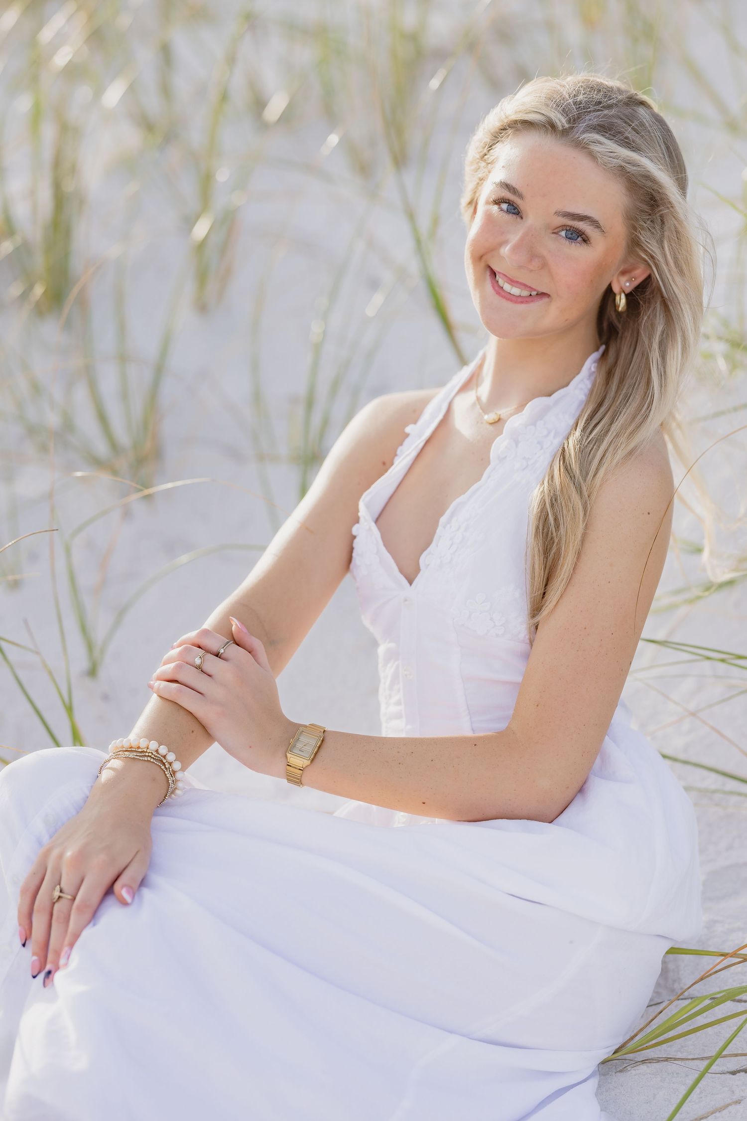 Smiling blonde woman in white halter dress sitting on sandy beach dunes with sea grass.