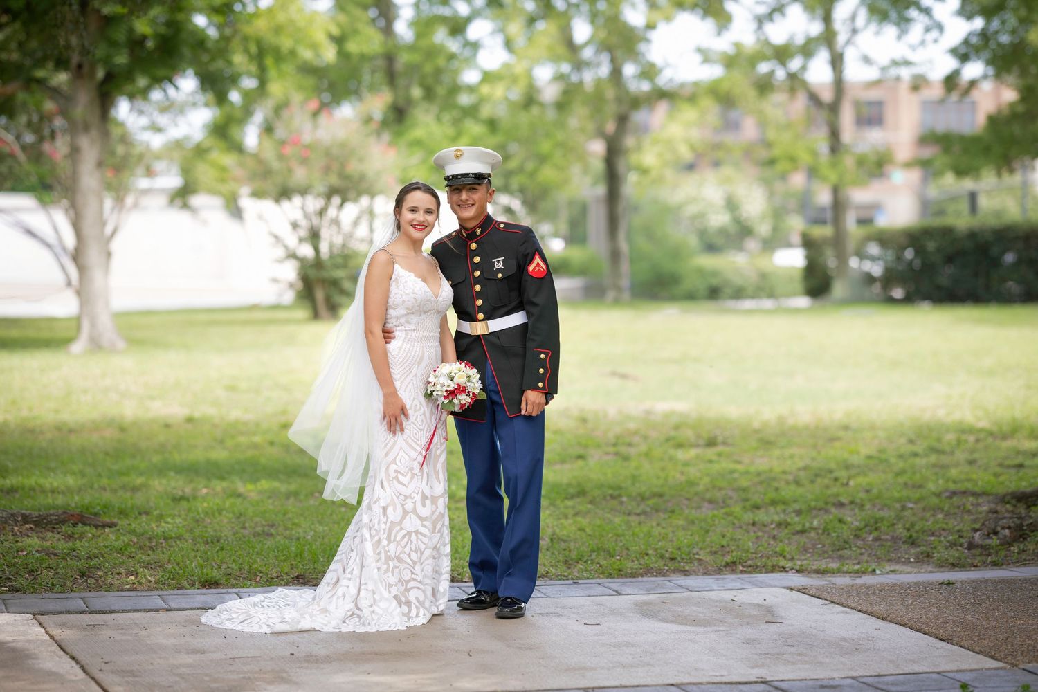 Bride in lace gown and groom in Marine dress uniform pose together outdoors on their wedding day.