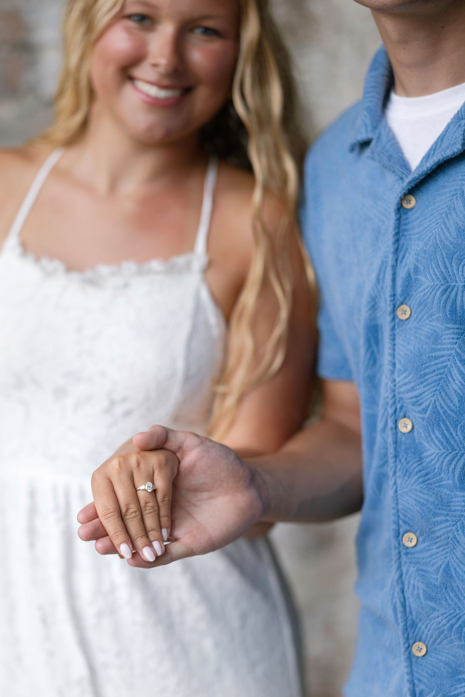 Smiling blonde woman in white dress showing off engagement ring while holding hands with man in blue shirt.