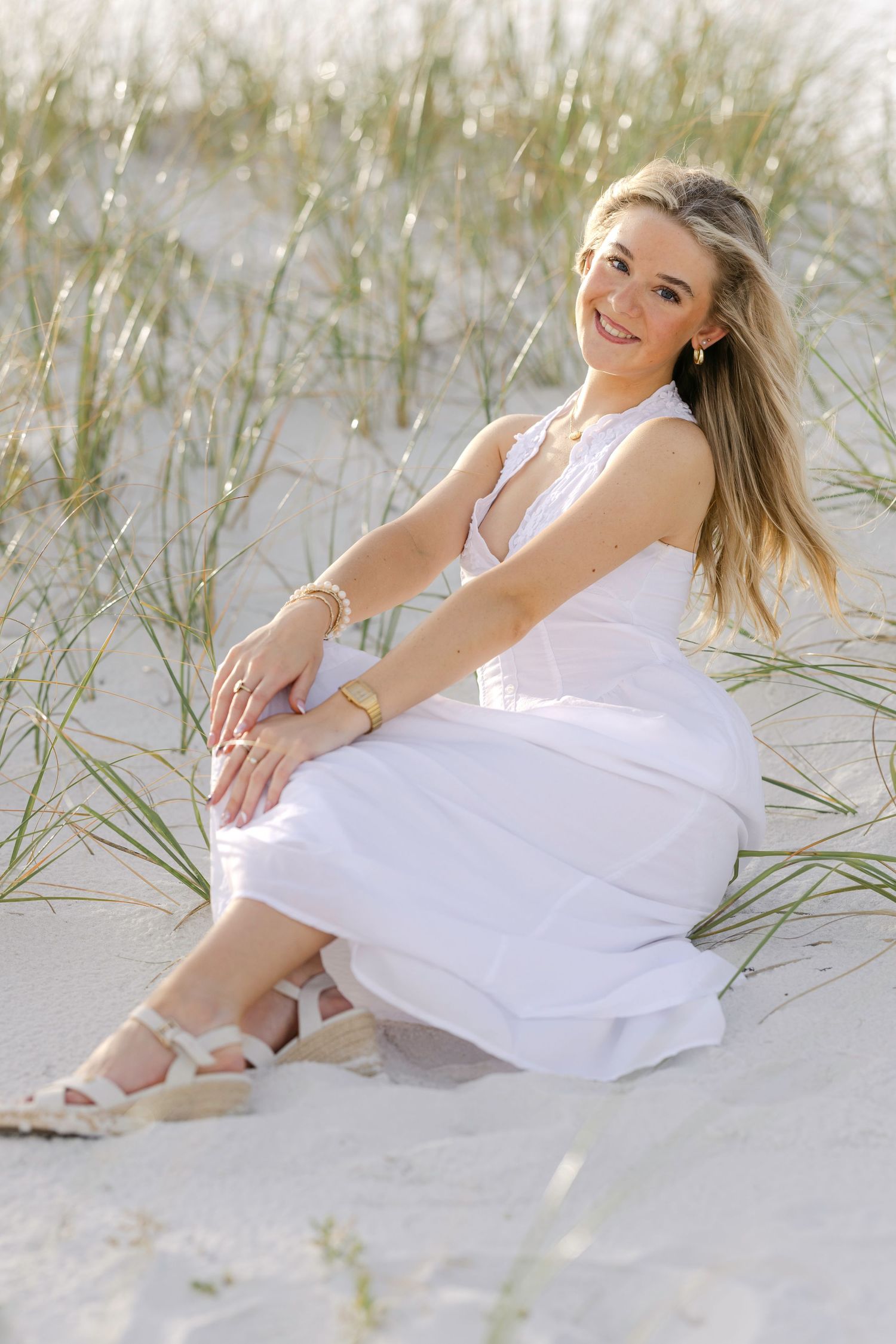 Young woman in white dress sitting on sandy beach dunes, smiling, with beach grass and soft sunlight.