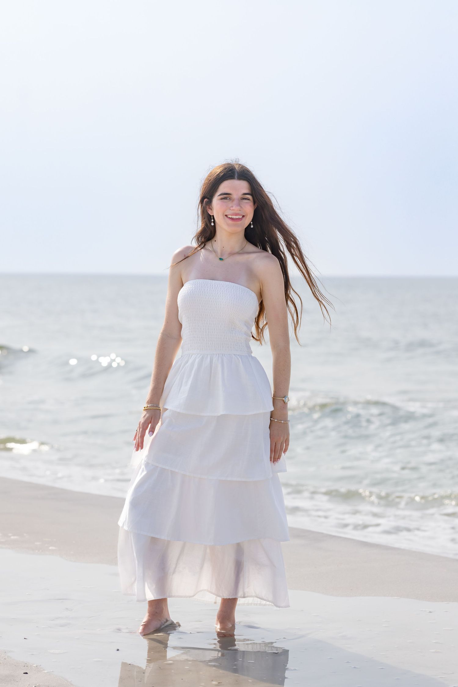 Young woman in white strapless tiered dress smiling on ocean beach with wind blowing her long hair.