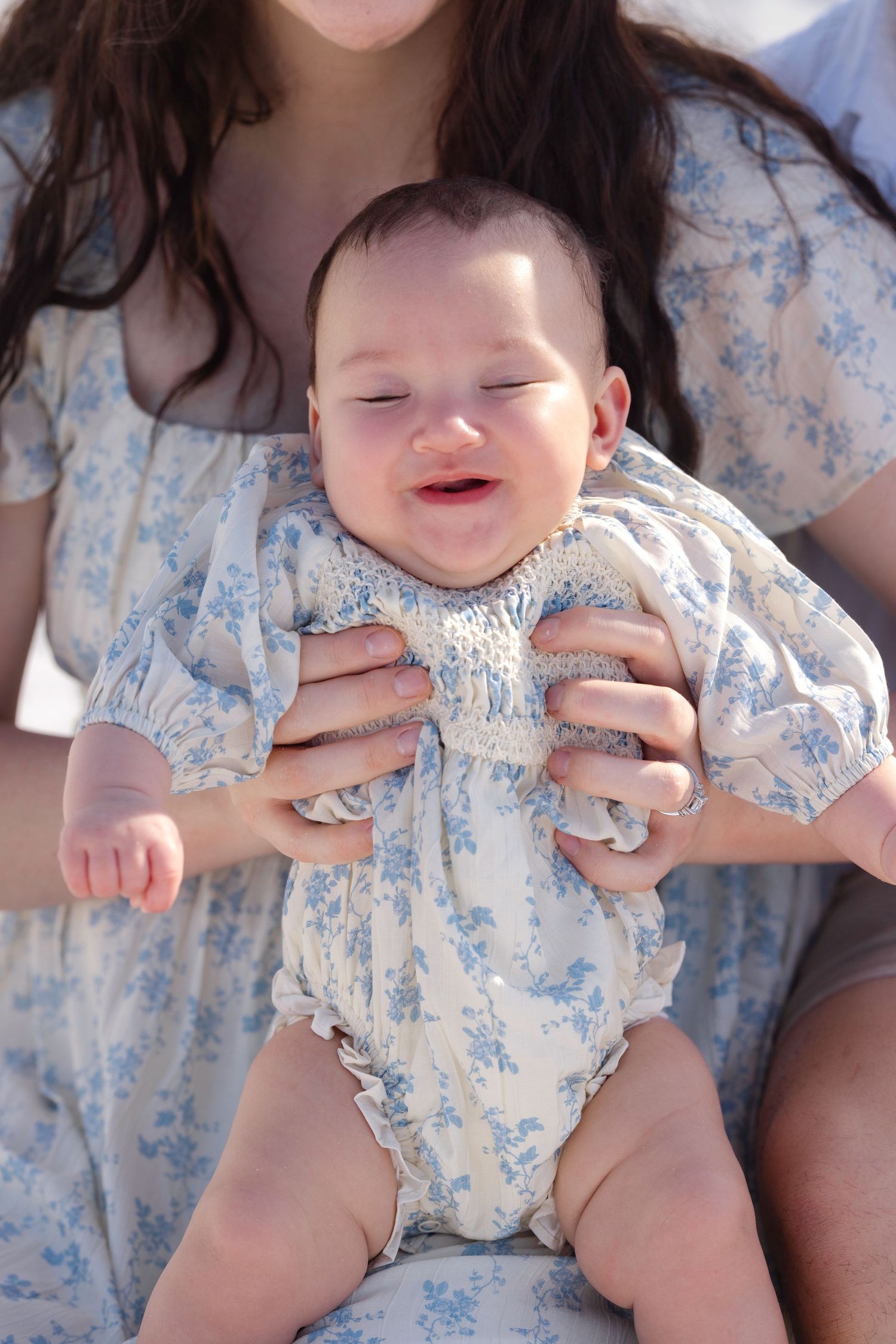Smiling baby in floral romper held by mother in matching blue floral dress outdoors.