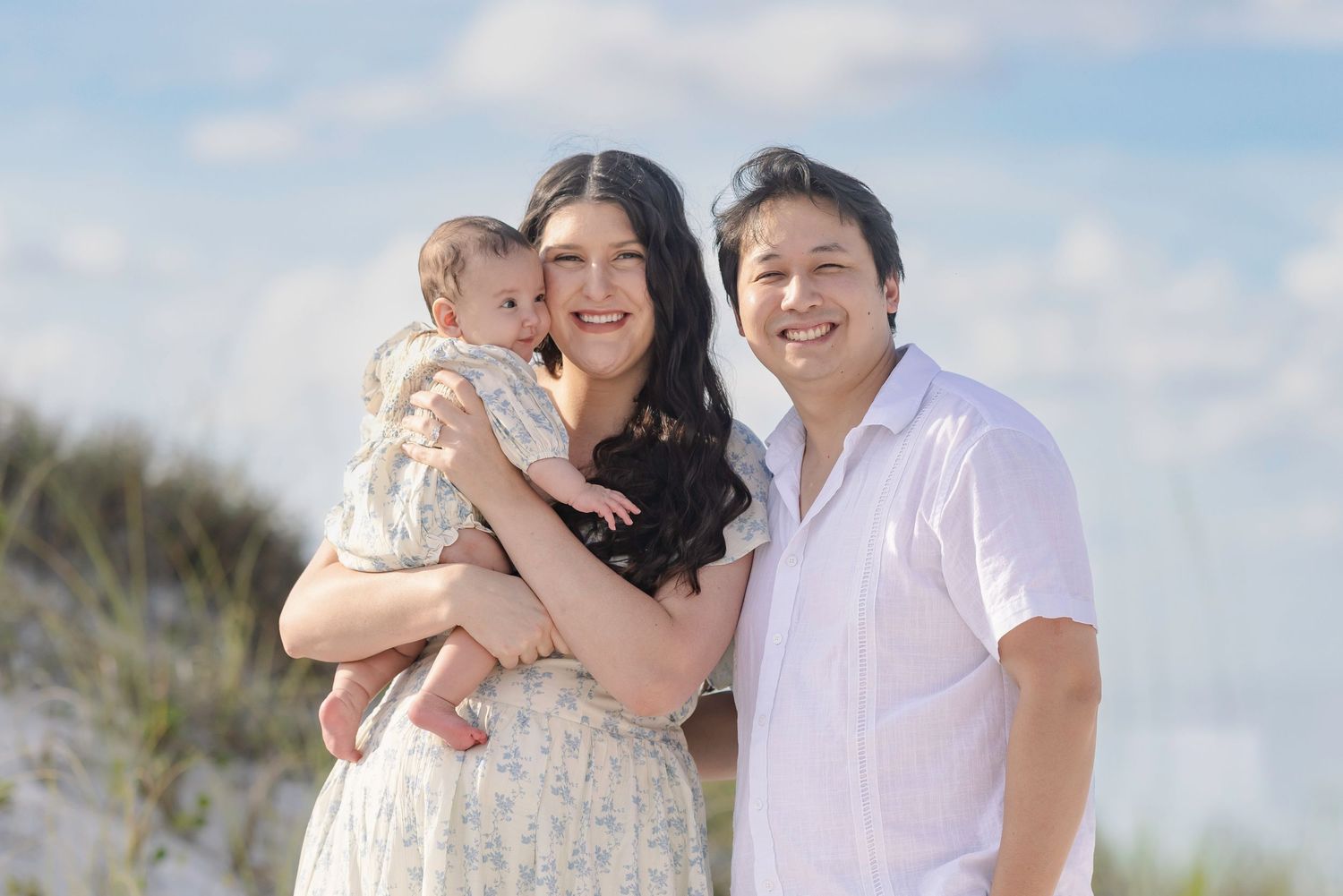 Happy family of three posing on a sandy beach, parents smiling while holding their baby on a sunny day.