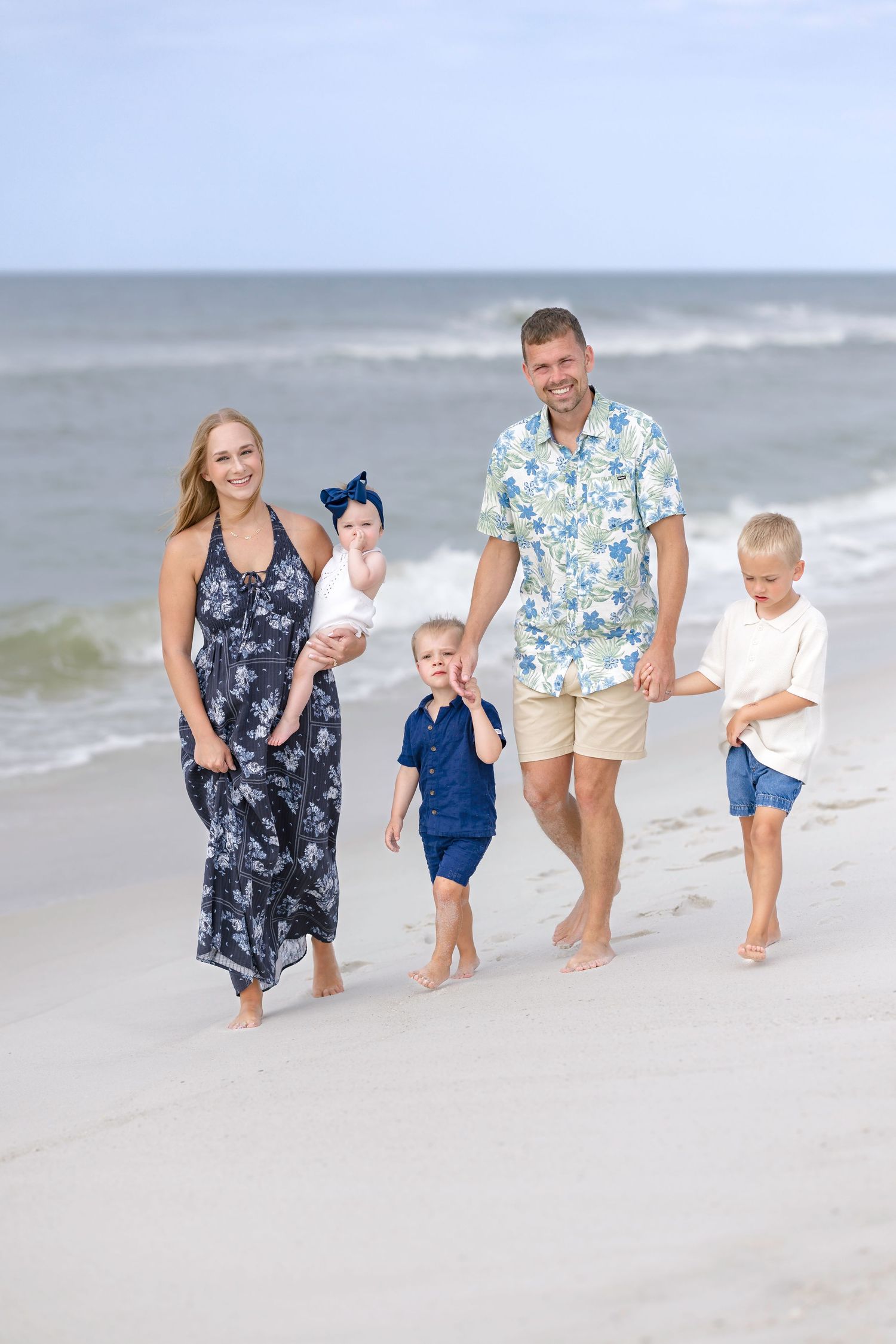 Happy family of five walking barefoot on beach shoreline, parents guiding three young children.