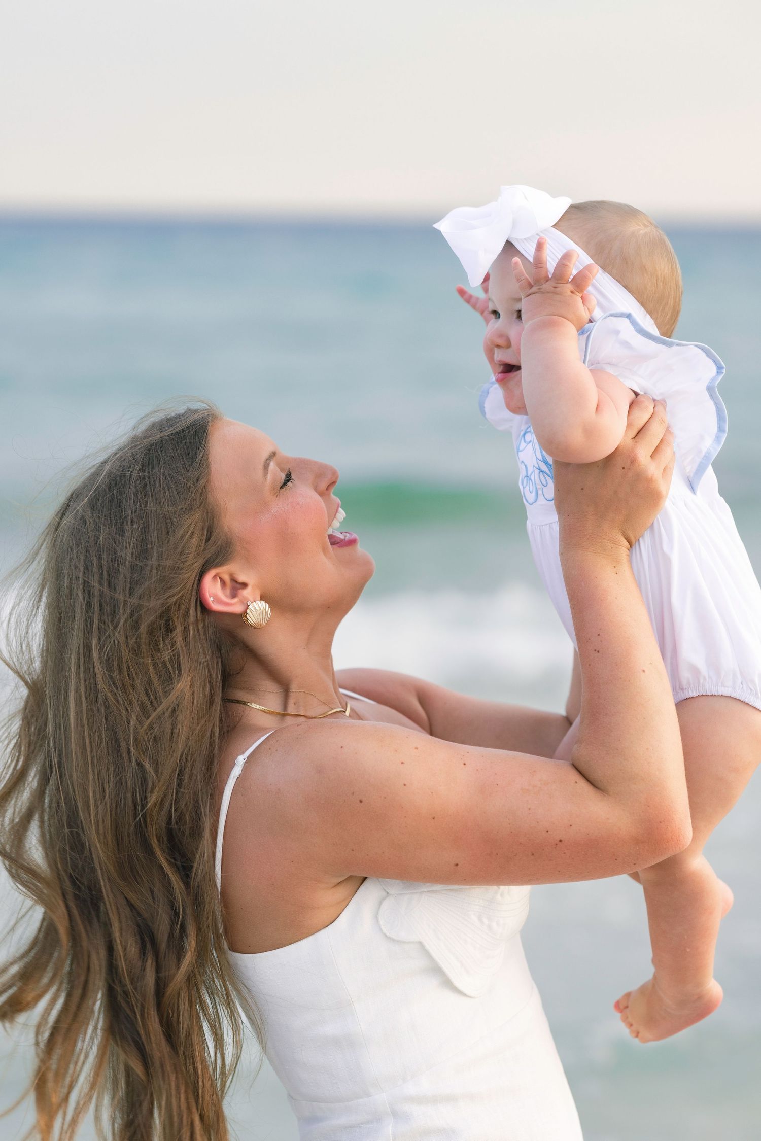 Young mother lifting joyful baby girl overhead with turquoise ocean waves in the background.