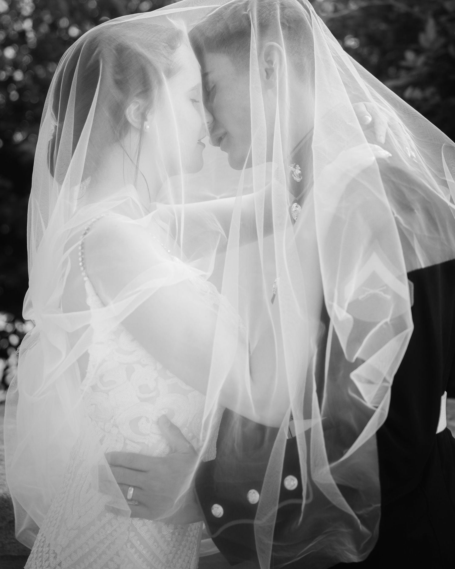 Black and white wedding photo of bride and groom sharing intimate moment under flowing veil.