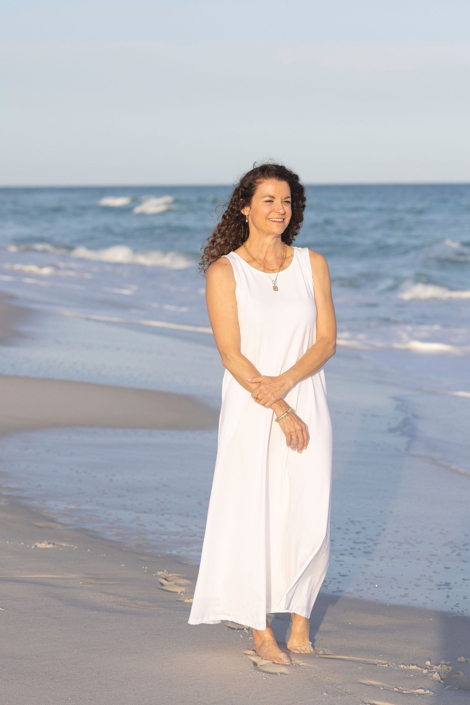 A smiling woman in a white maxi dress stands on a sandy beach with ocean waves in the background.