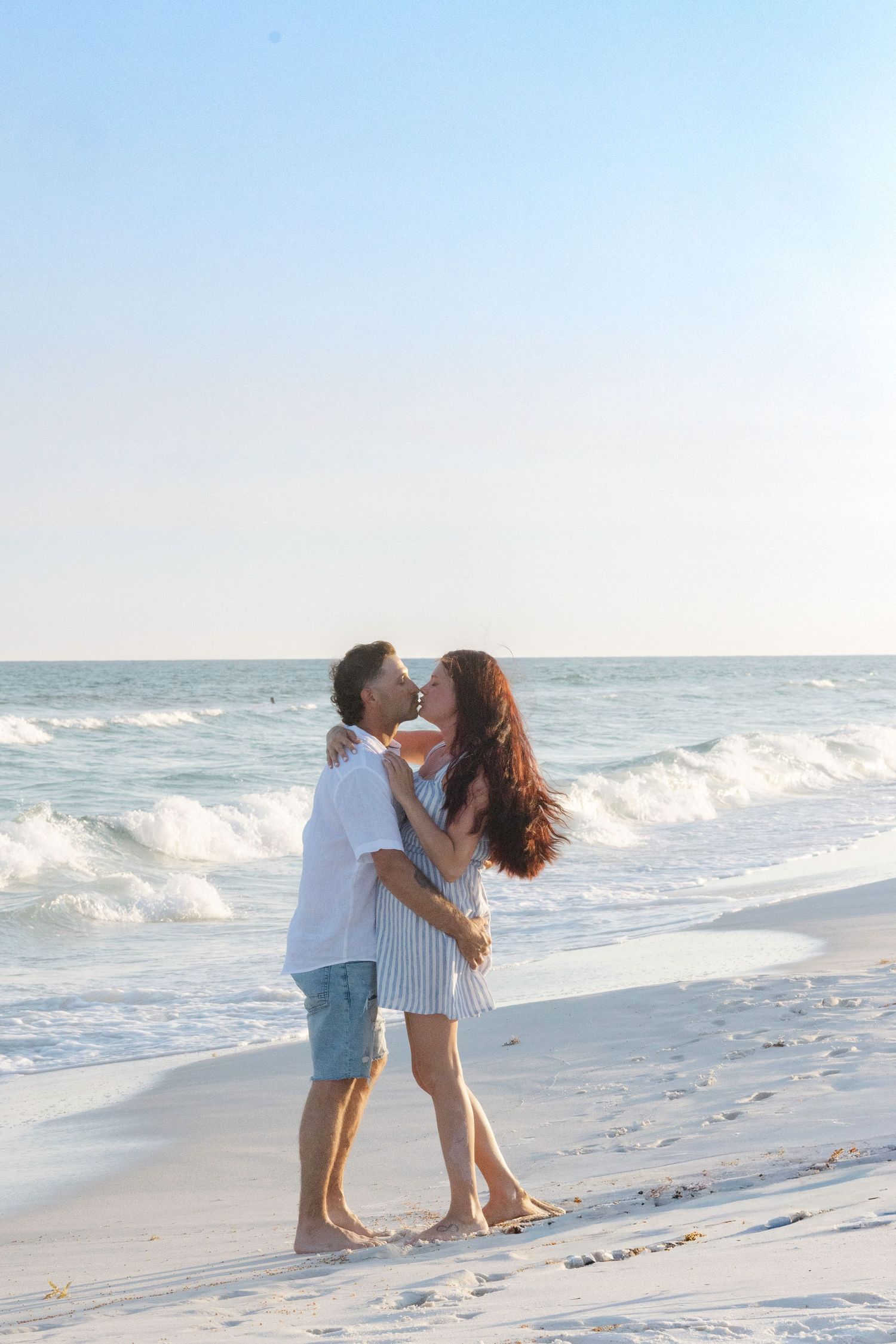 A couple shares a romantic kiss on a white sand beach with ocean waves behind them at sunset.