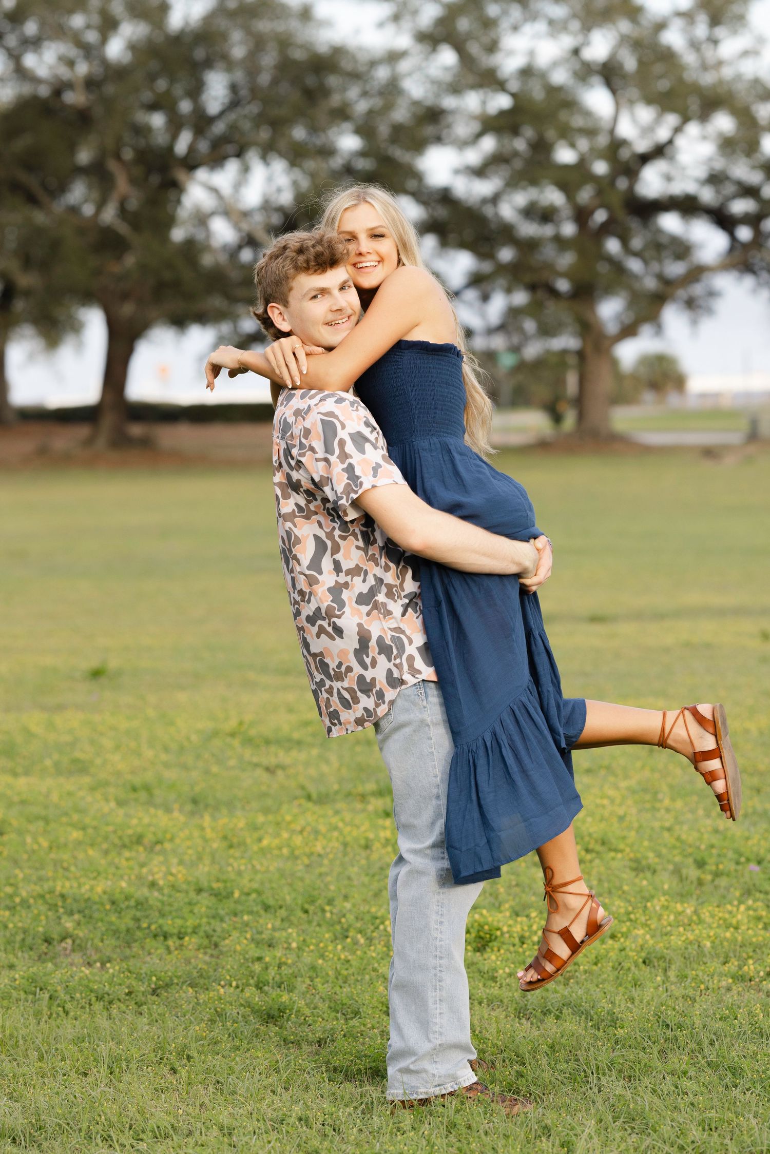 Couple embraces joyfully in a grassy field on a sunny day wearing casual outfits and brown sandals.