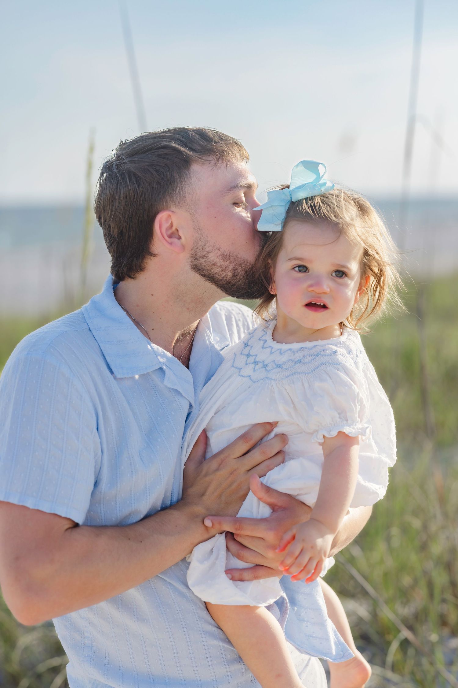 A bearded father kisses his toddler daughter with a blue bow on a sunny beach with sea oats behind them.