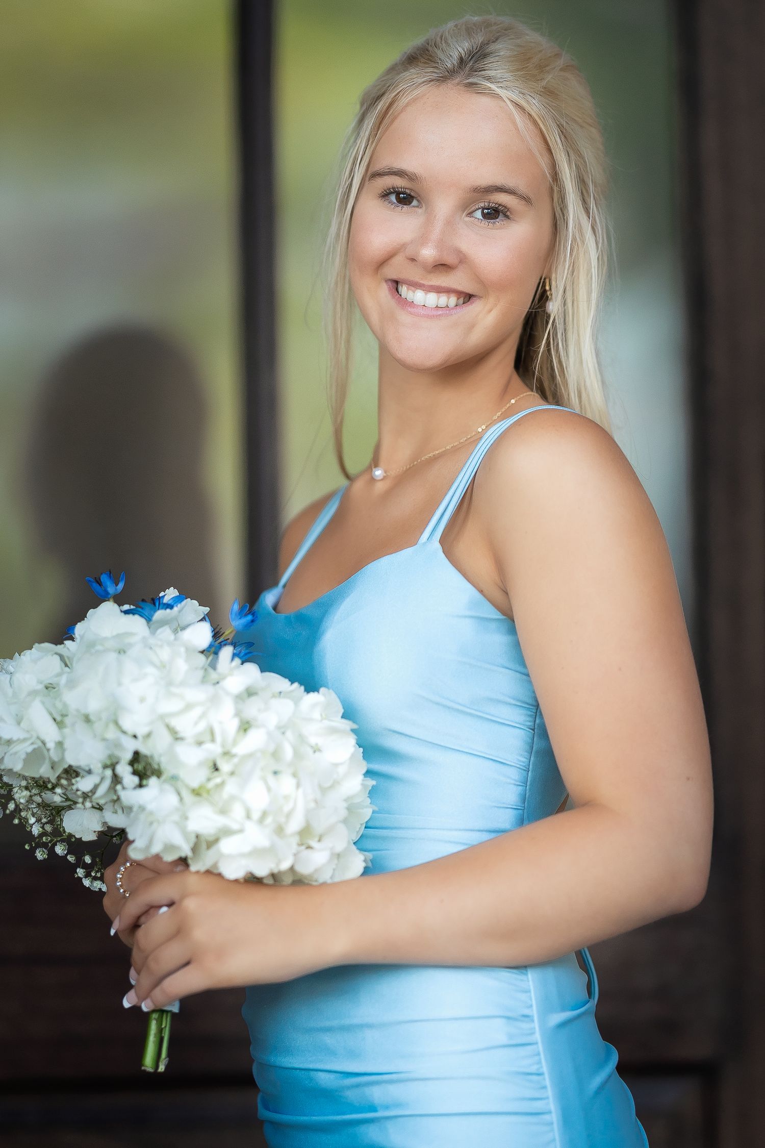 Smiling blonde bridesmaid in blue satin dress holding white hydrangea bouquet at wedding ceremony.