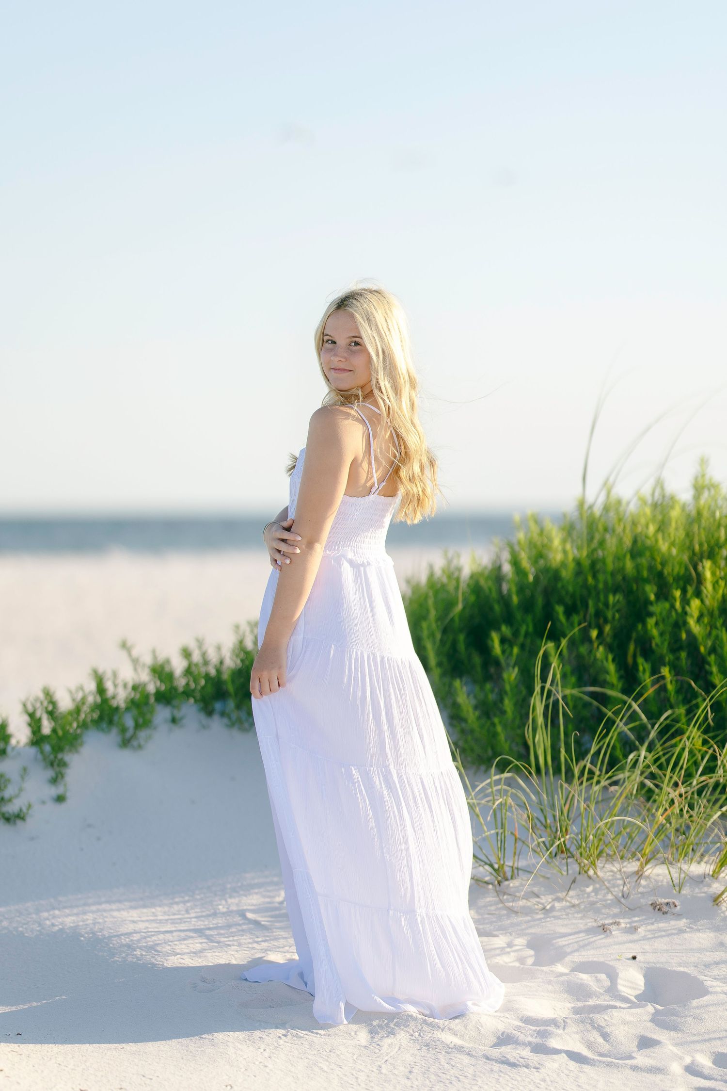 Blonde woman in white maxi dress standing on sandy beach dunes with ocean in background.