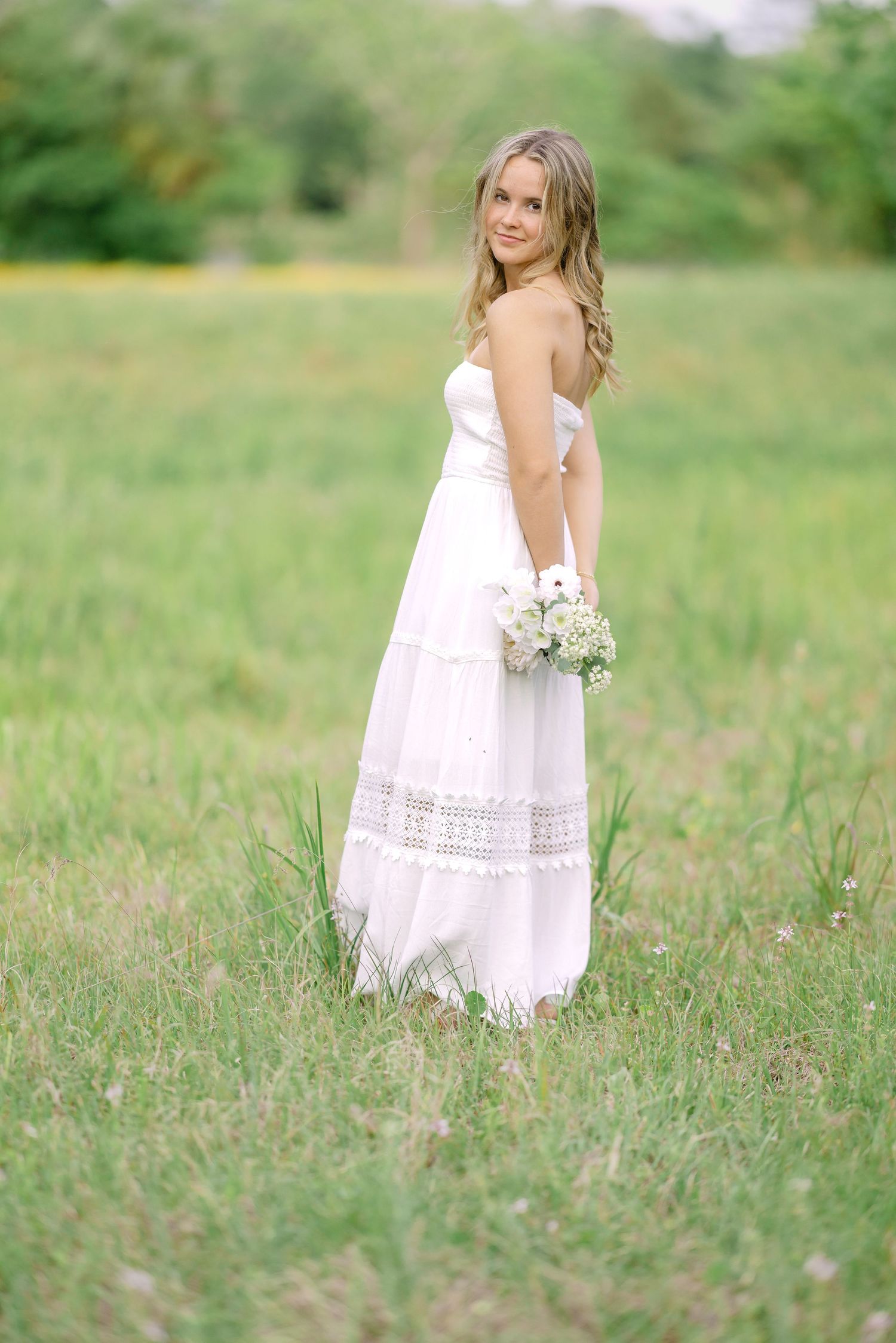 Young blonde woman in white strapless gown holding small bouquet stands in green meadow, looking back over shoulder.